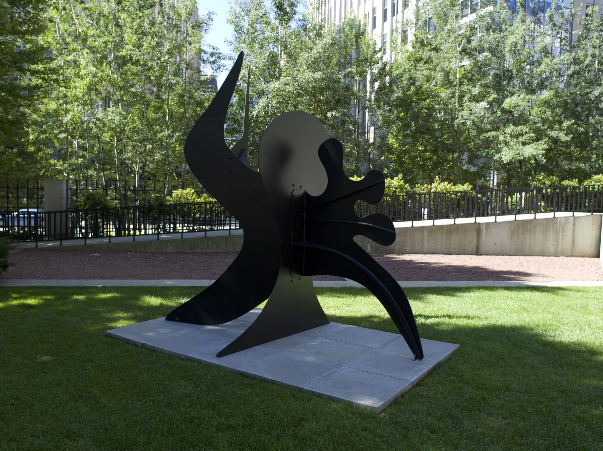 A three-dimensional, brown, abstract sculpture with silhouetted shapes is placed on a concrete slab within a park.