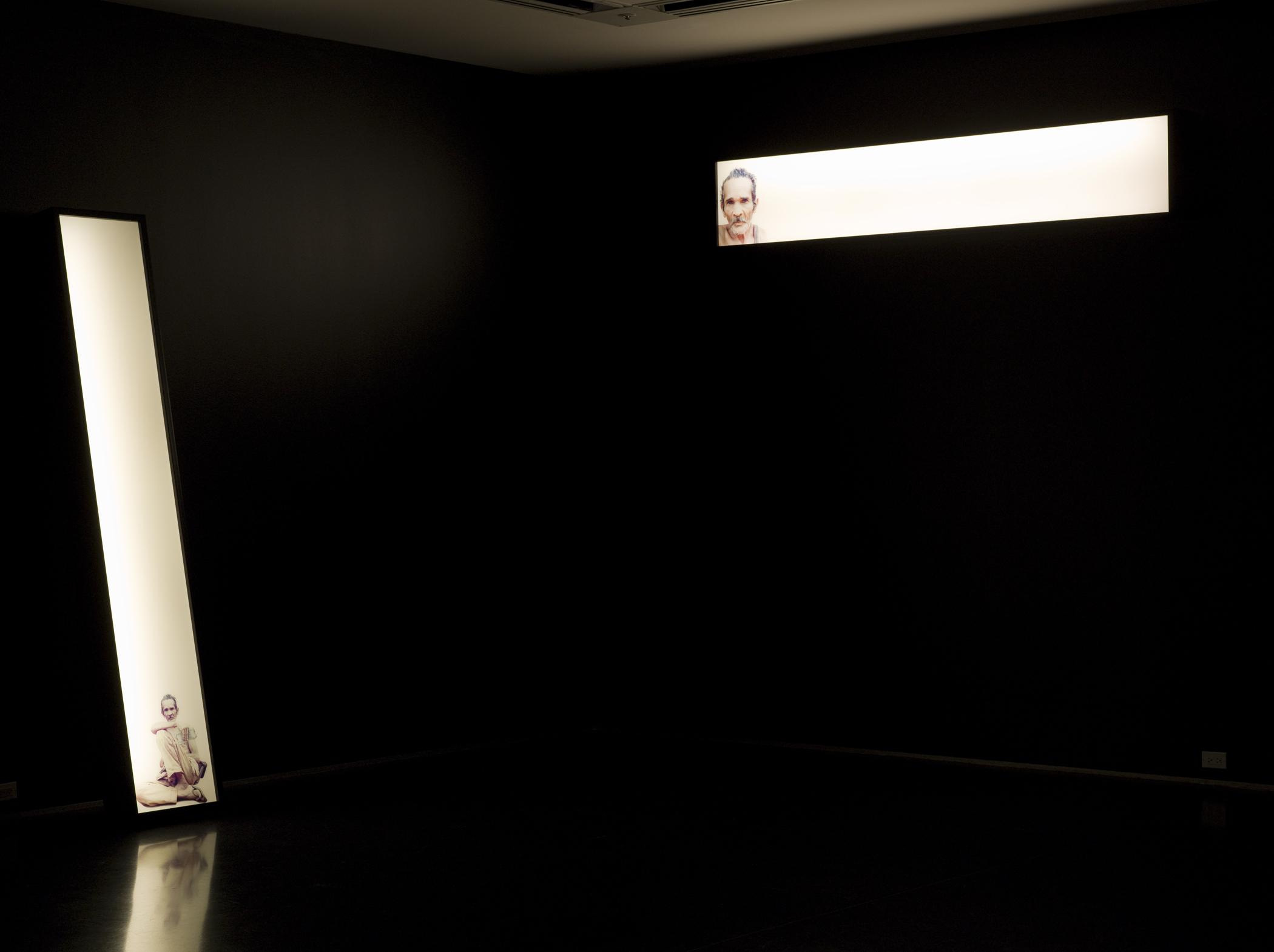Two narrow white glowing rectangles depicting a man illuminate a pitch-black room. The horizontal box on the right hangs high on the wall; the other leans against the wall to the left.