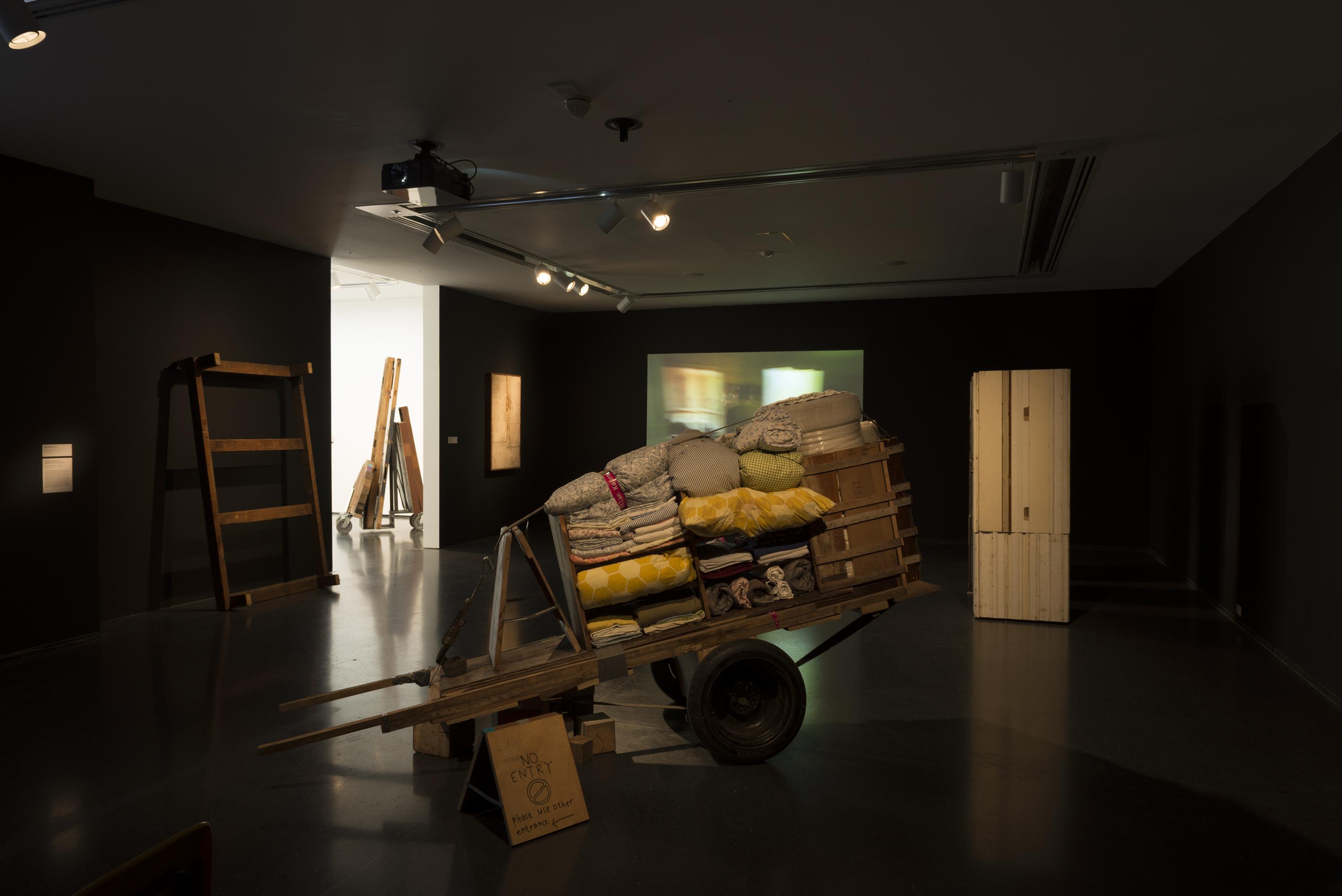 The setting is a dark room with black walls. In the middle is a makeshift wooden two-wheeled hand cart loaded with yellow and grey folded fabrics. On the back wall, a video is playing.
