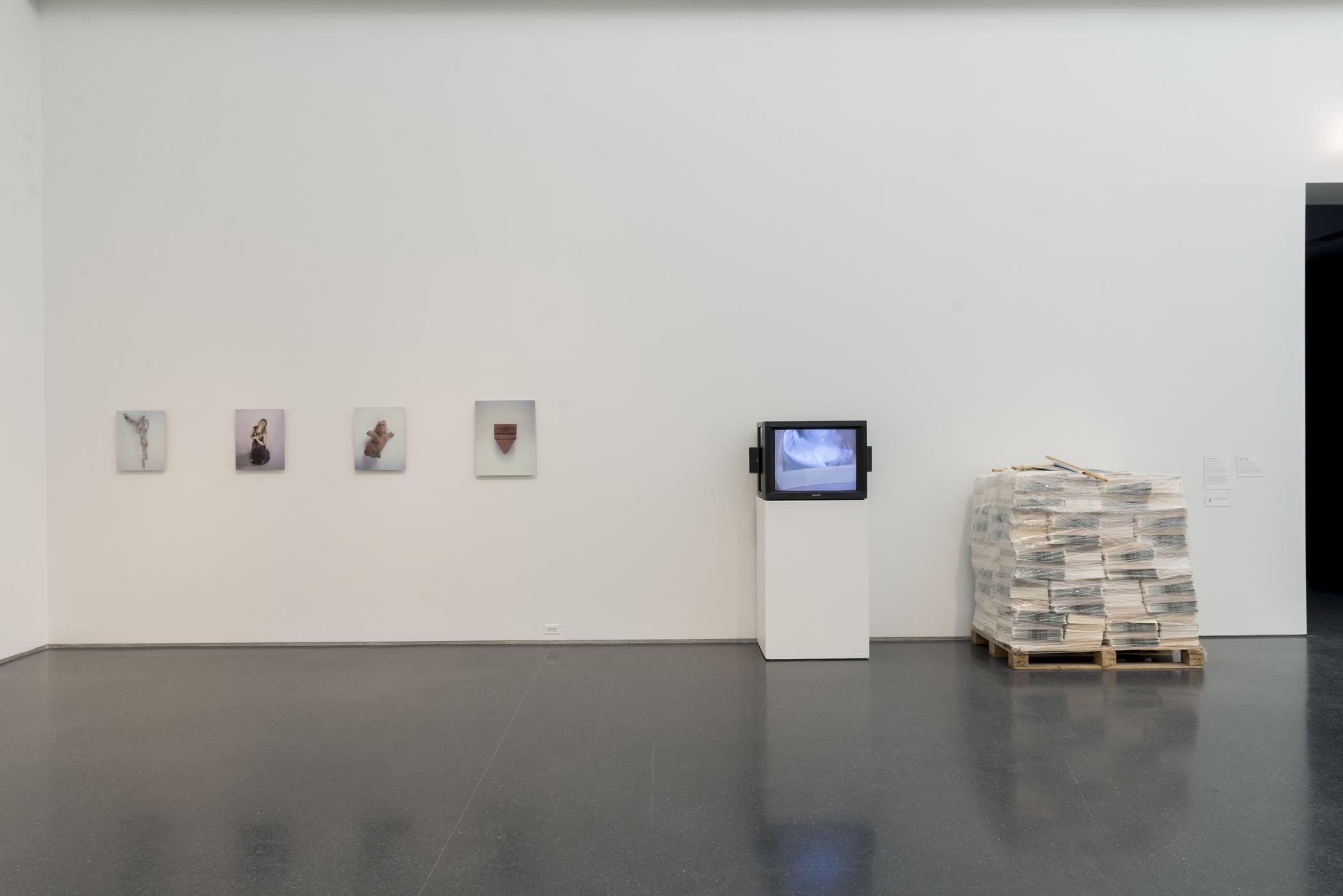 Four photographs of ancient sculptures hang on a white wall. A podium with a television playing a video and a wooden platform that has multiple mounds of paper on it sit adjacent to the photographs, against the wall.