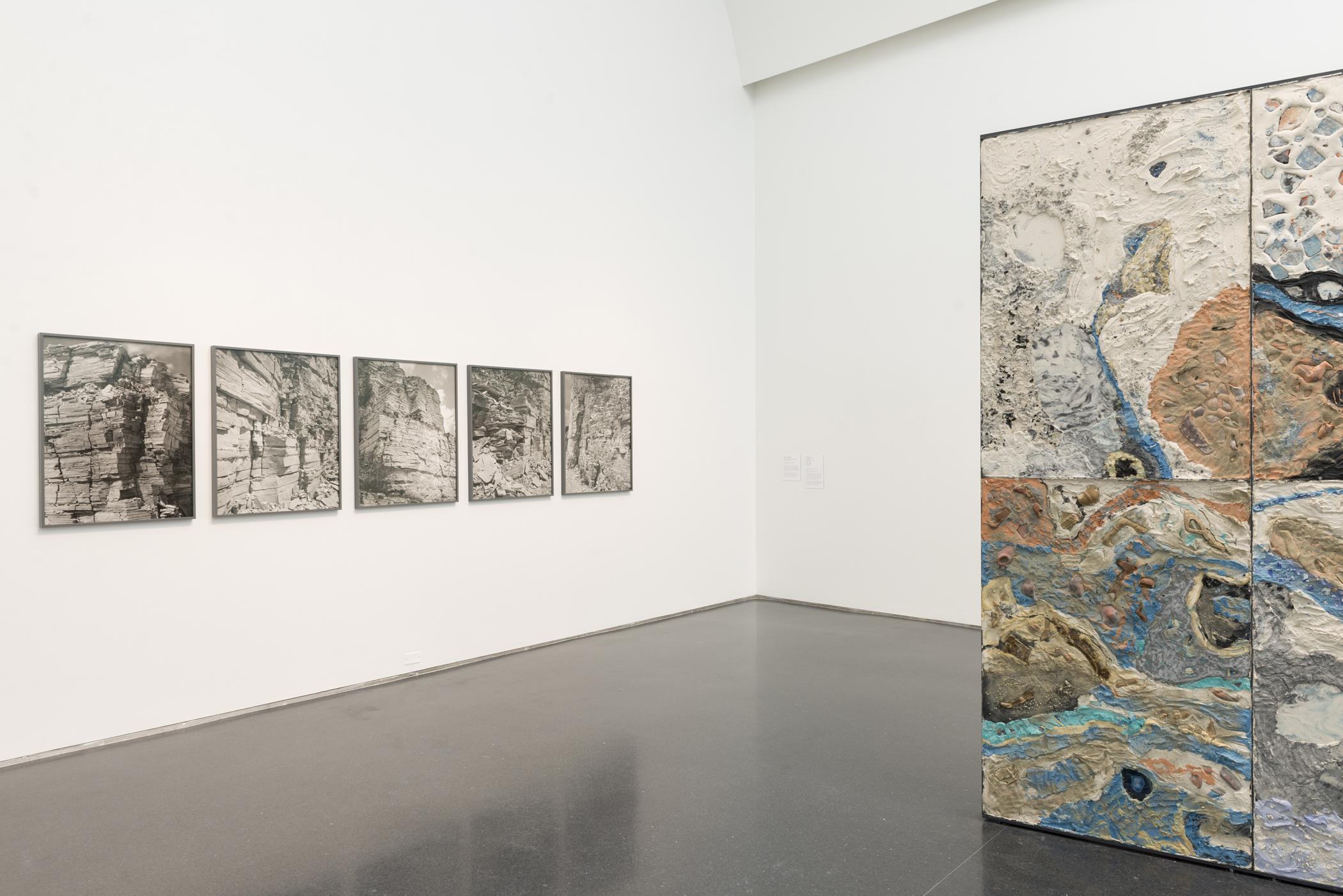 Four photographs are hung on a white wall to the left of a rectangular sculpture. The photographs are pictures of rock ledges in black and white. The rectangular sculpture is in blue and earth tones resembling the texture and look of a rock ledge.