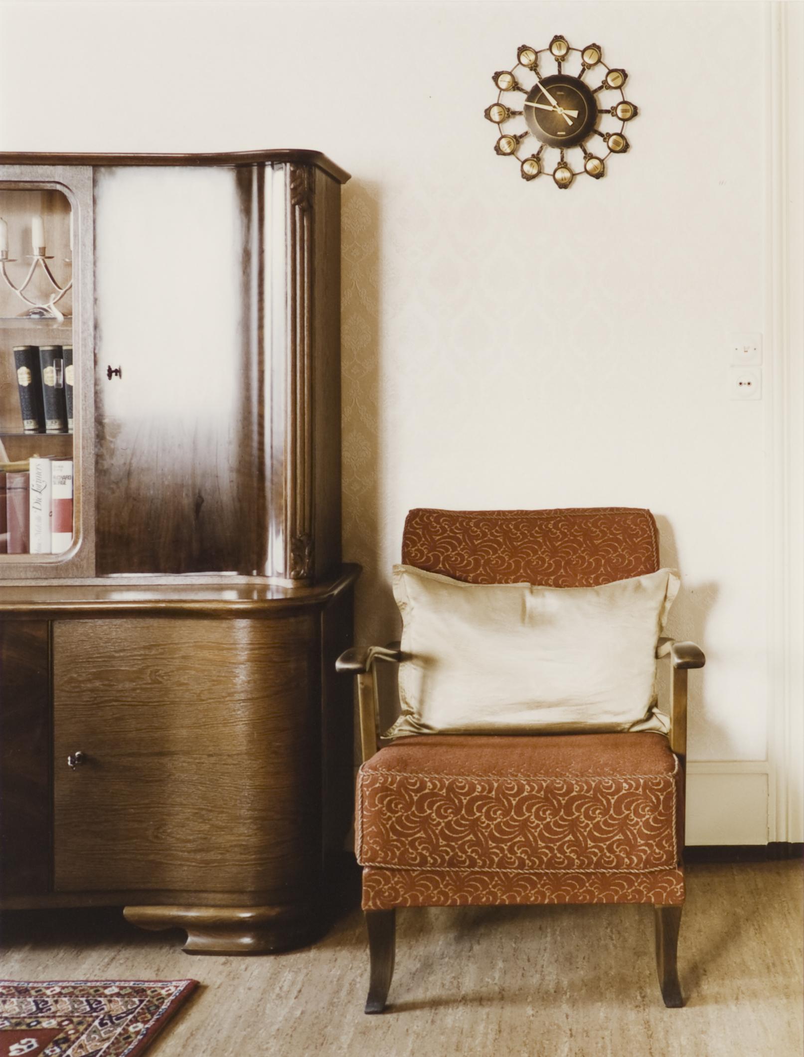 A sepia-toned image shows an armchair with a wooden bureau to the left of it and a clock hung on the wall.