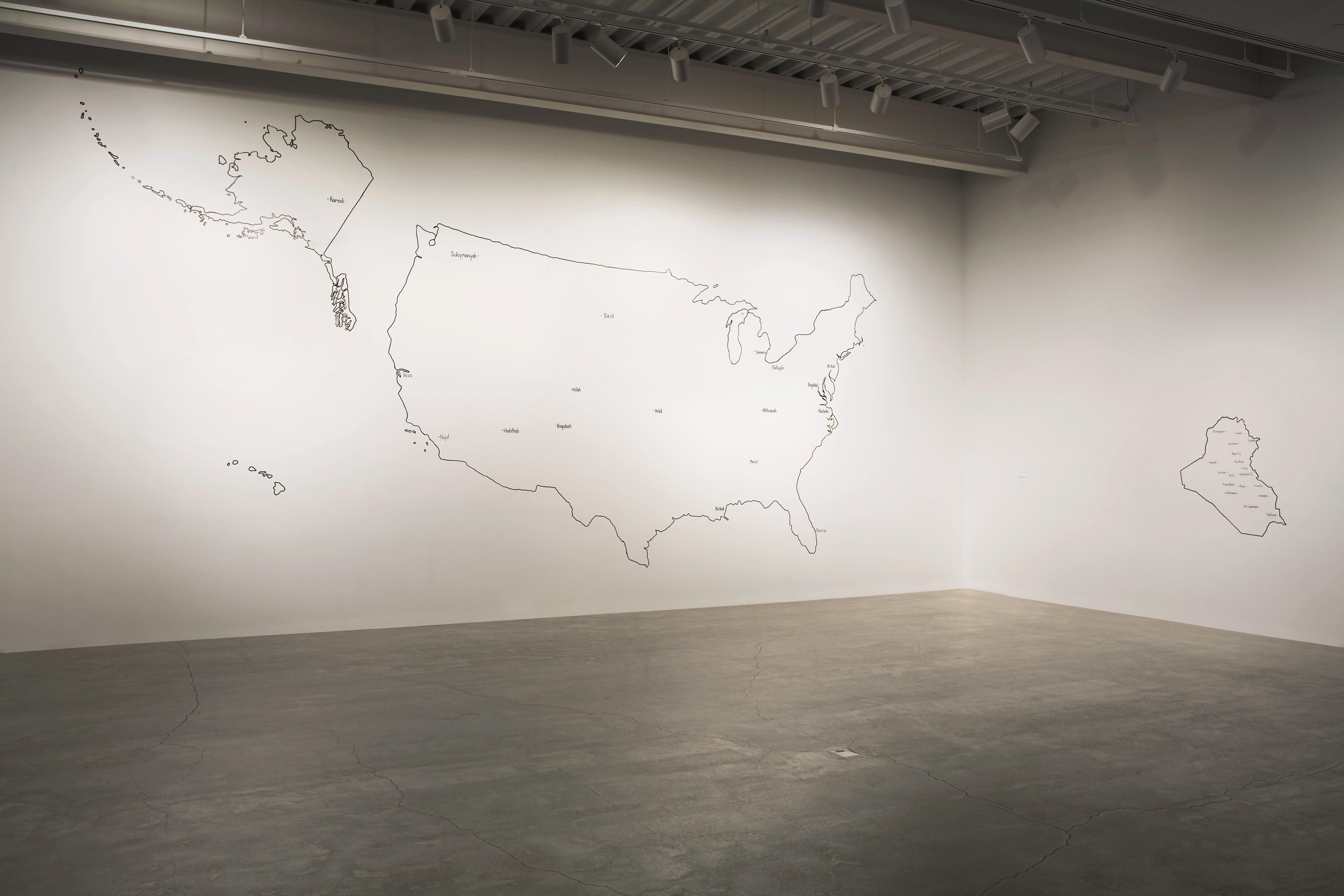 On a vast white wall, a map of the United States is outlined in black while a map of Iraq is outlined in black on a large white wall to its left.