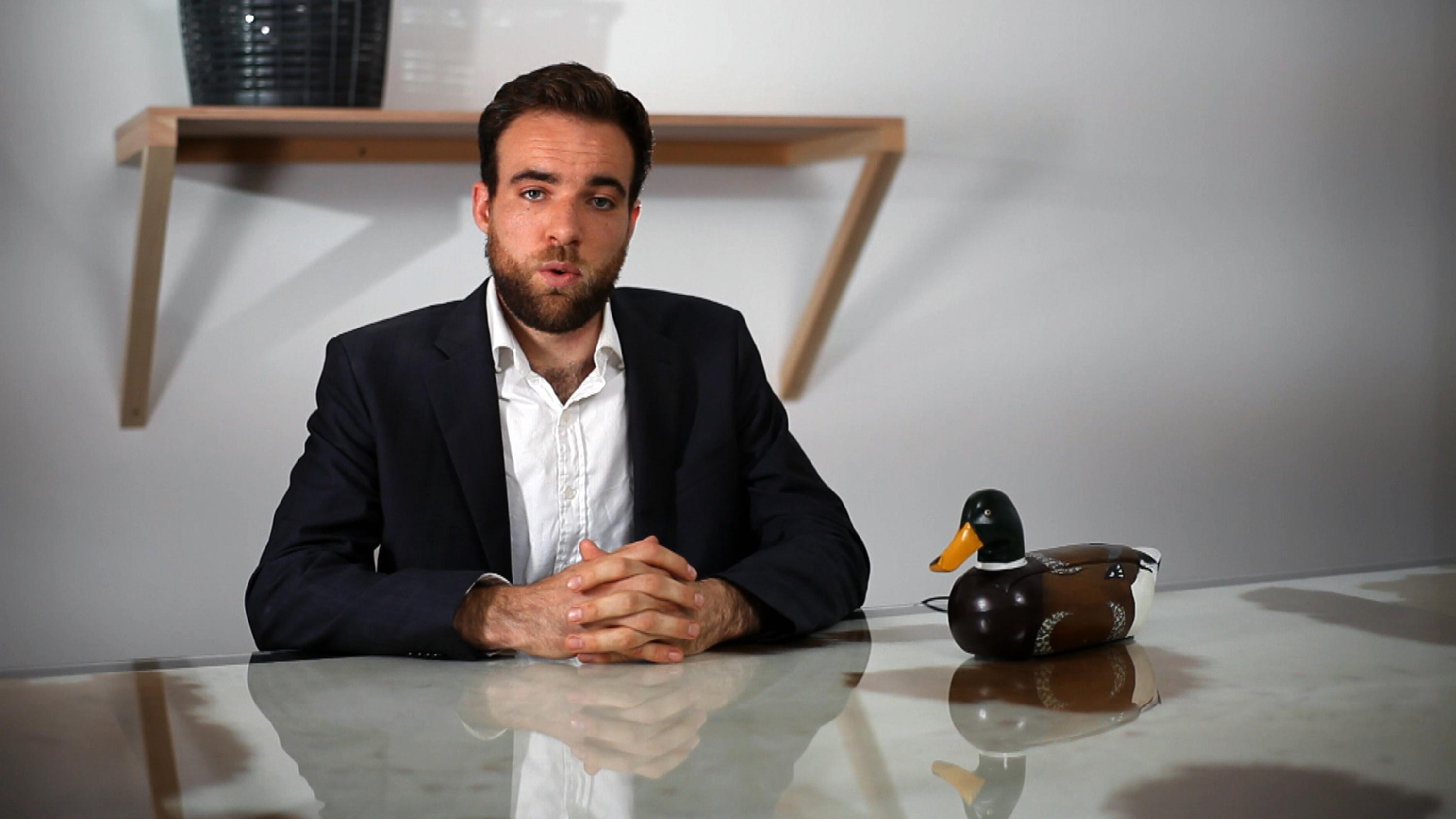 A man in a suit jacket sits at a table with hands folded in front of him and a duck phone to his left. His lips are pursed, as if in mid sentence.