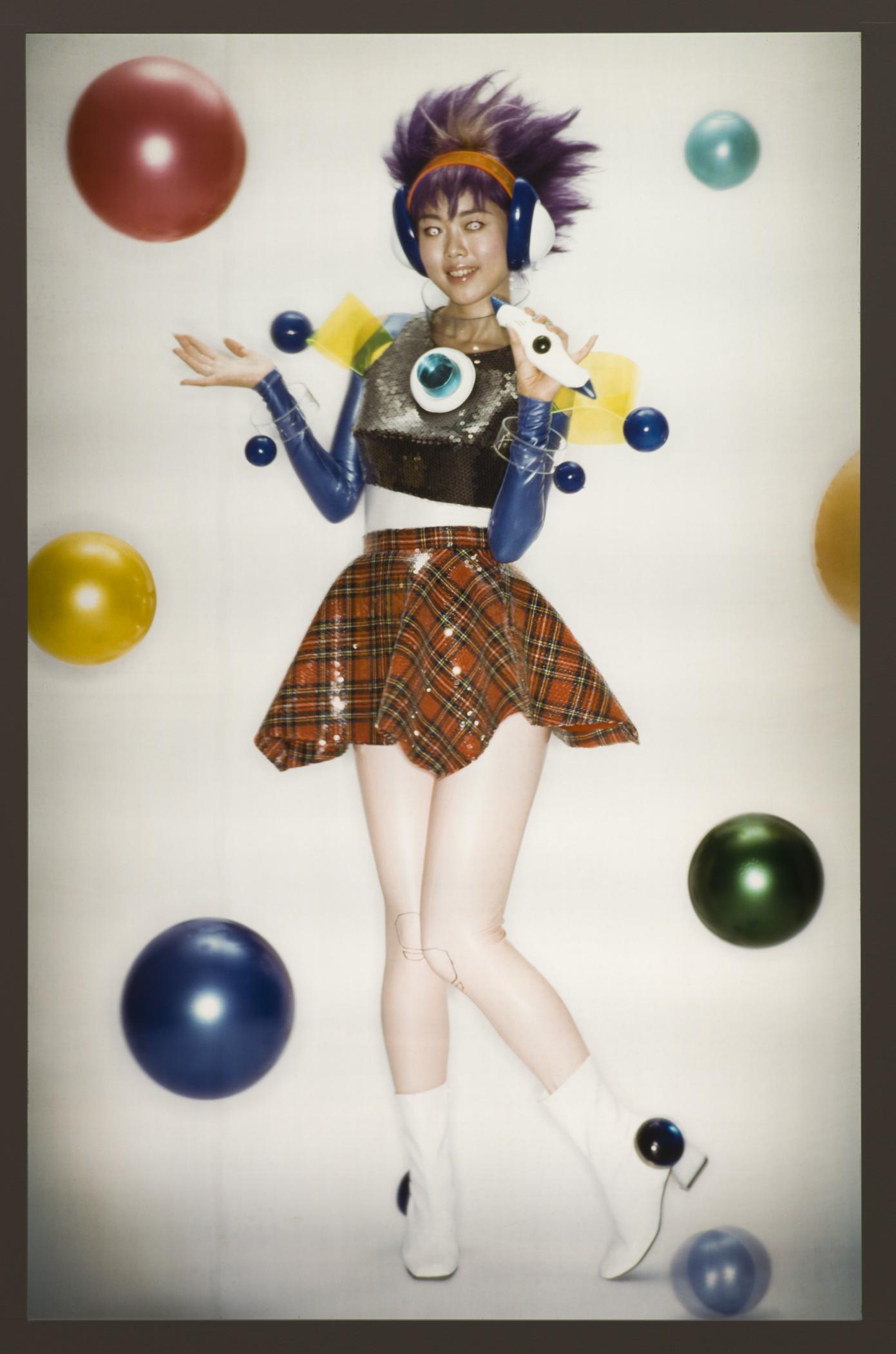 A futuristic person wearing a brightly colored crop top and a skirt poses in between thirteen colorful spheres.