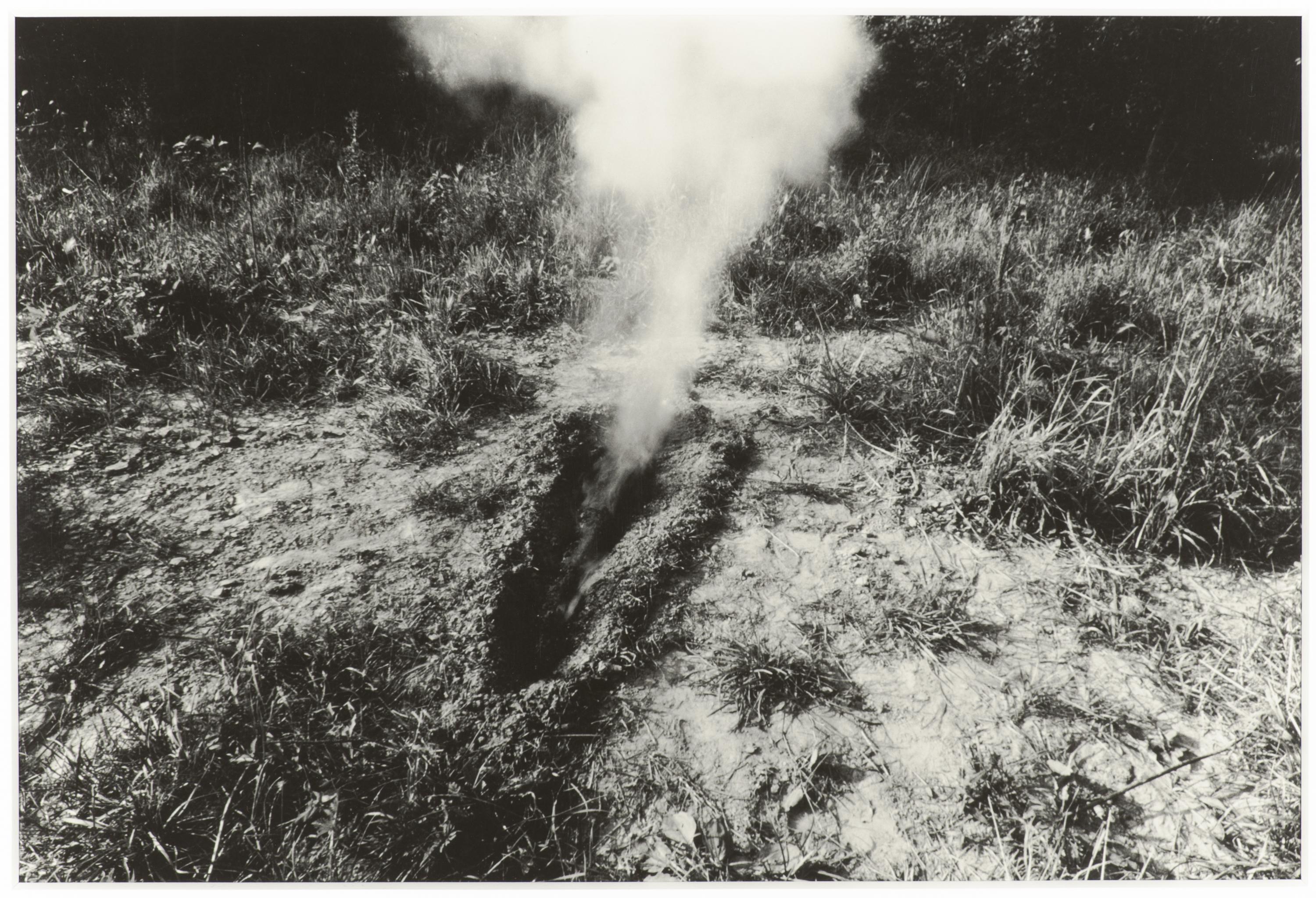 A black-and-white photograph depicts smoke billowing from a body-sized trench in a clearing with weeds and trees on the horizon.