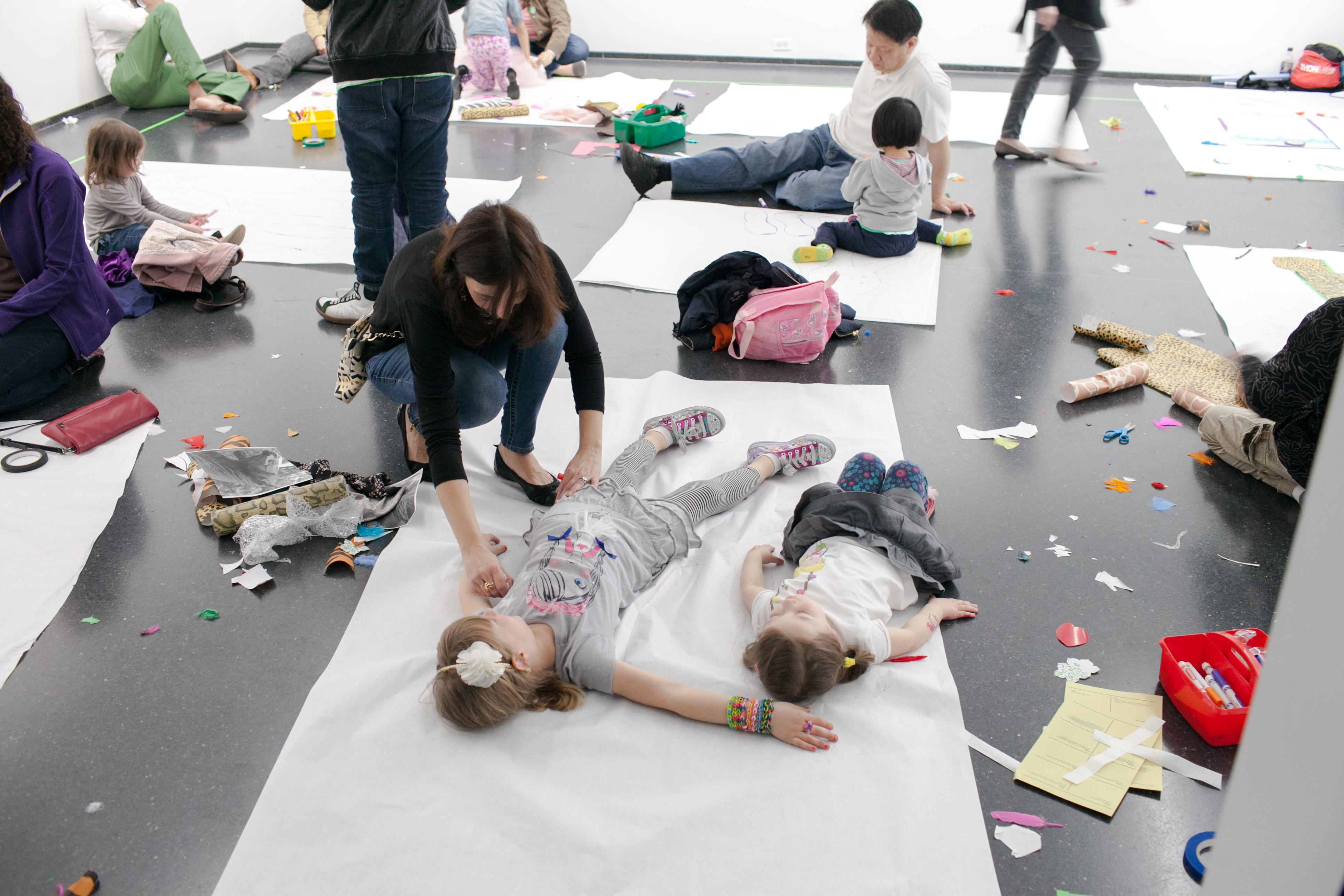 Parents and children interacting together in a white-walled gallery, with children lying down on large sheets of white craft paper while their parents trace the outline of their bodies.