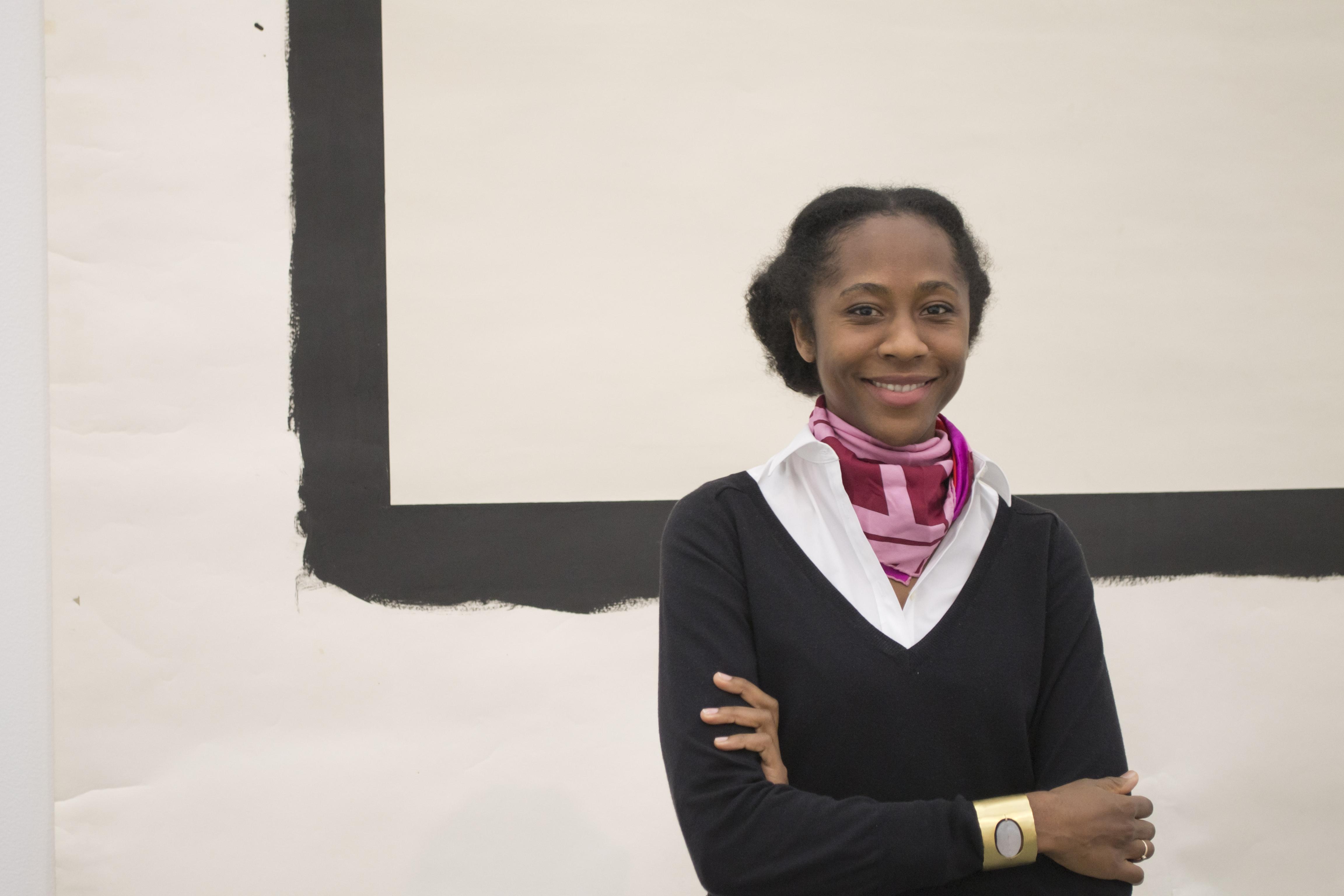 A woman in a black sweater and pink scarf smiles in front of back and white artwork.