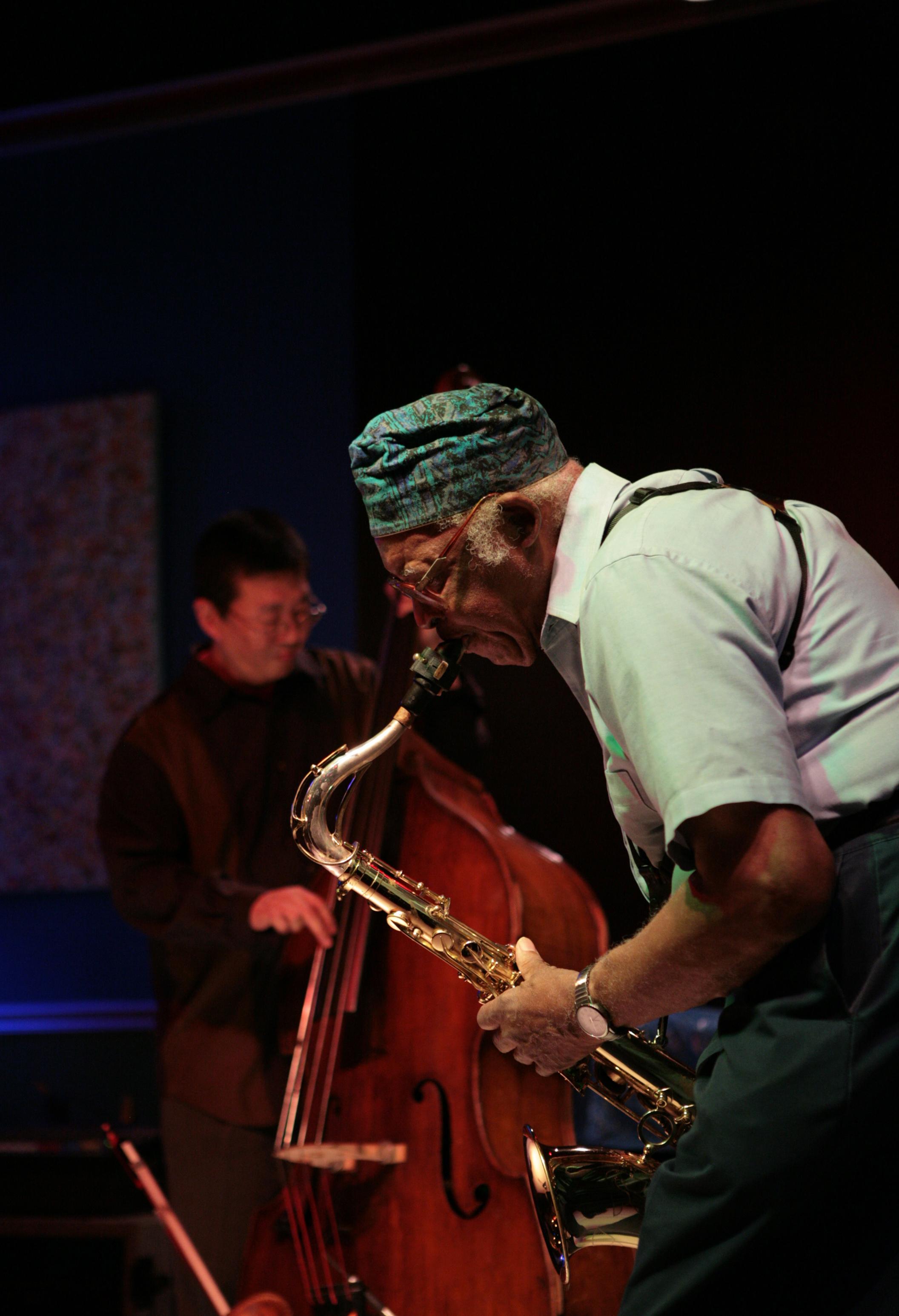 A person wearing a patterned green and blue cap bends forward playing a saxophone, while another person plays standing bass in the background.