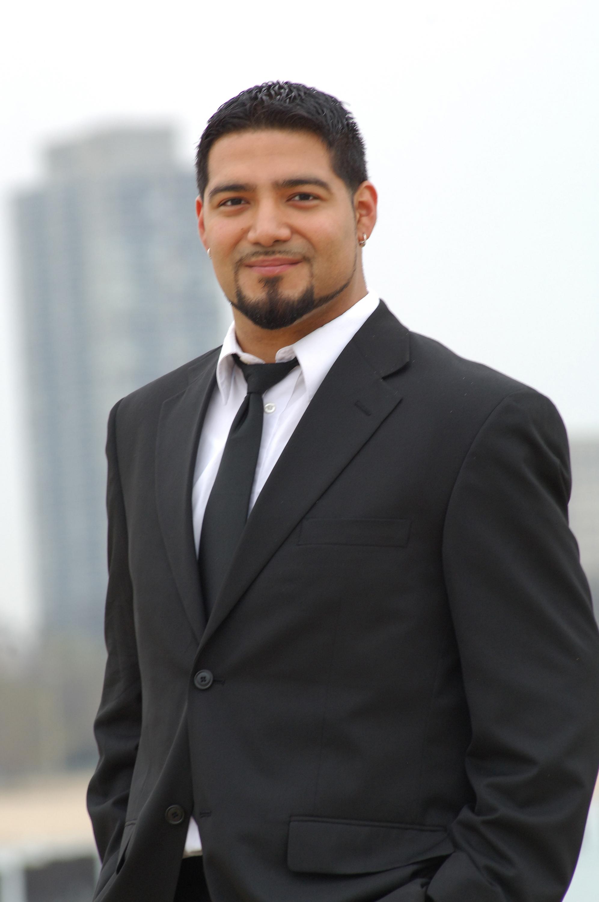 A portrait of a tan-skinned man with short dark hair and a groomed goatee in a black suit and tie, stands outside with skyscrapers blurred in the distance.