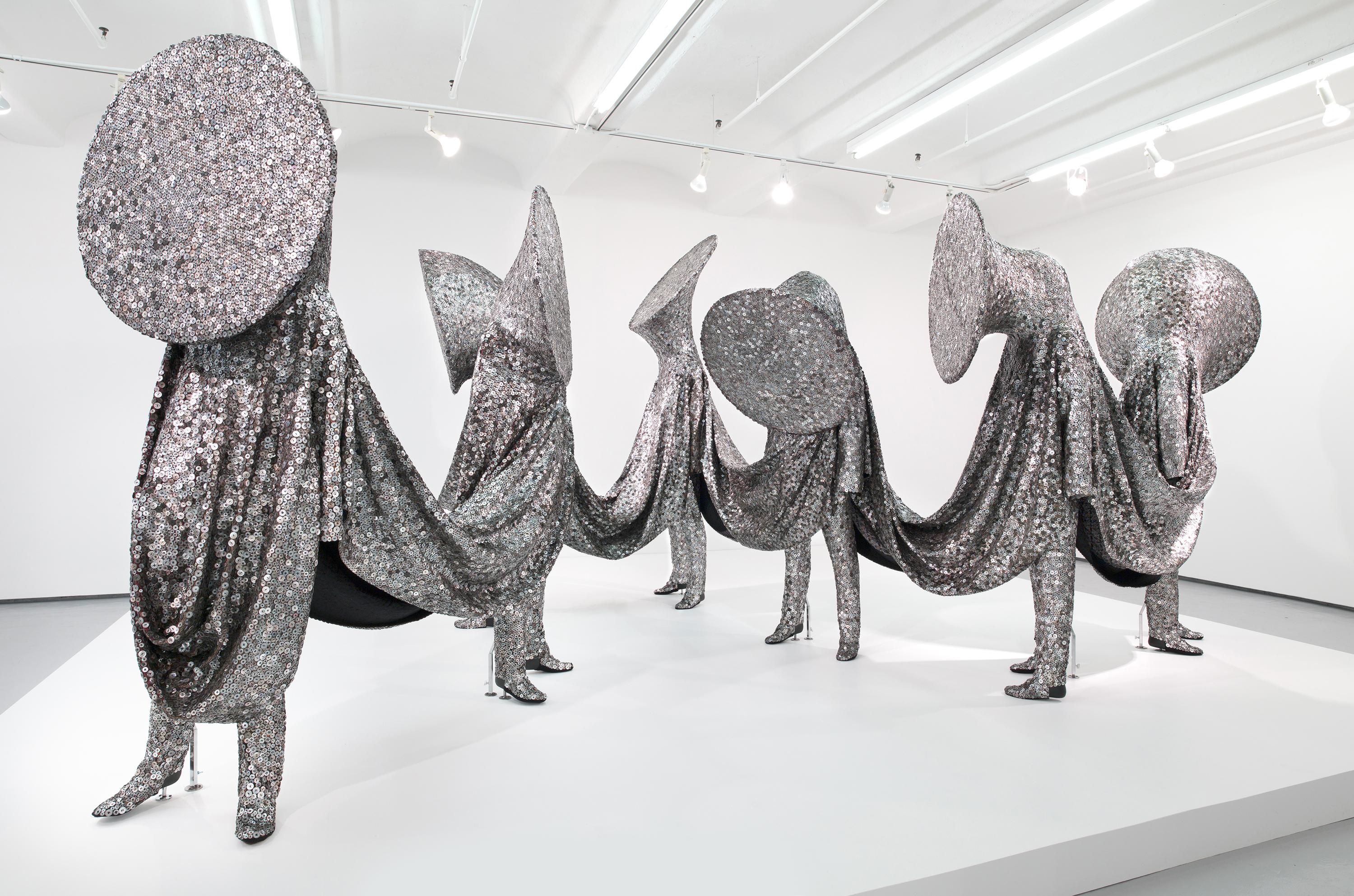 Seven human figures with brass instrument heads are entirely covered in a shiny silver sequined fabric that connects each of them.