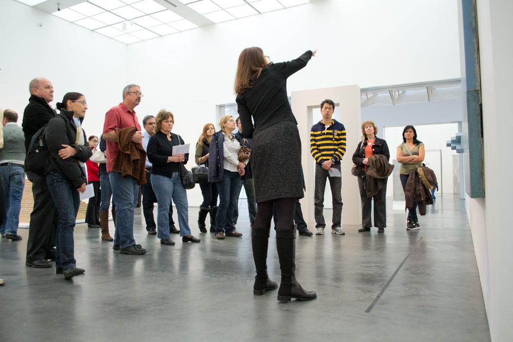 A woman points out a detail in an artwork as a group of people surround her listening and looking at the artwork.