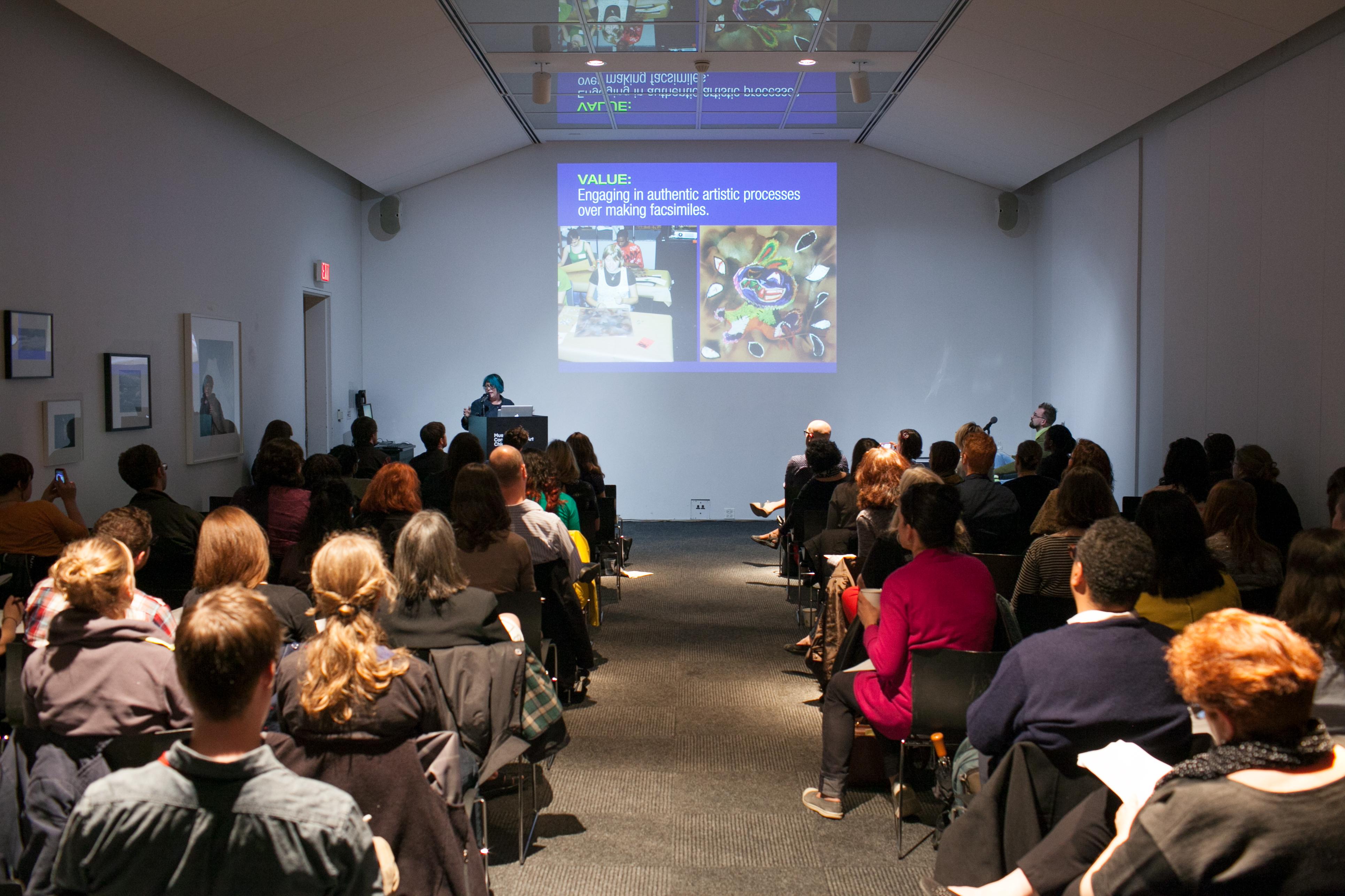 A room is full of people sitting in rows of chairs, taking notes, and looking up at a projection of a presentation, while someone speaks at the front podium.