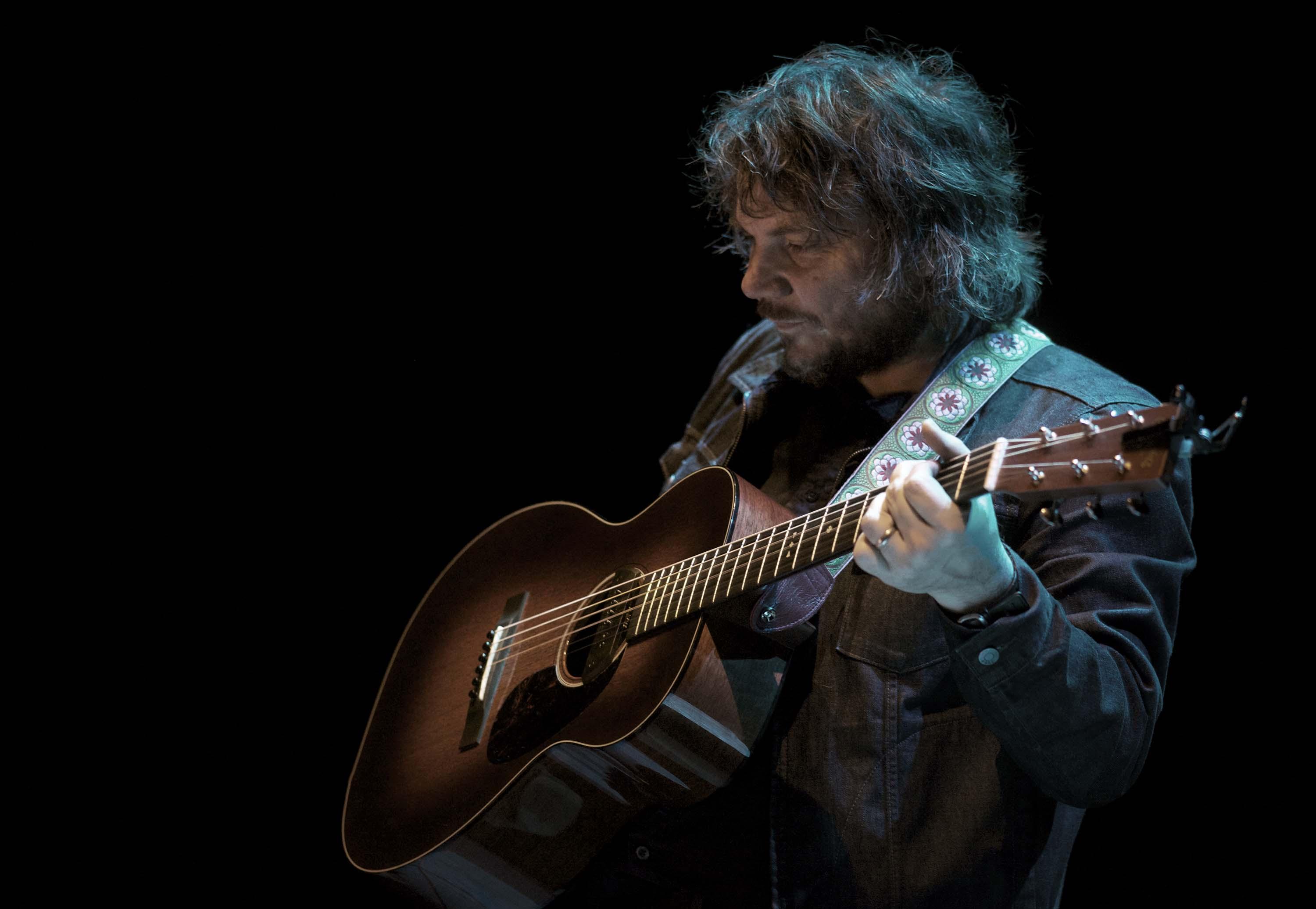 A man with long shaggy hair wearing a black denim jacket holds a wooden acoustic guitar.