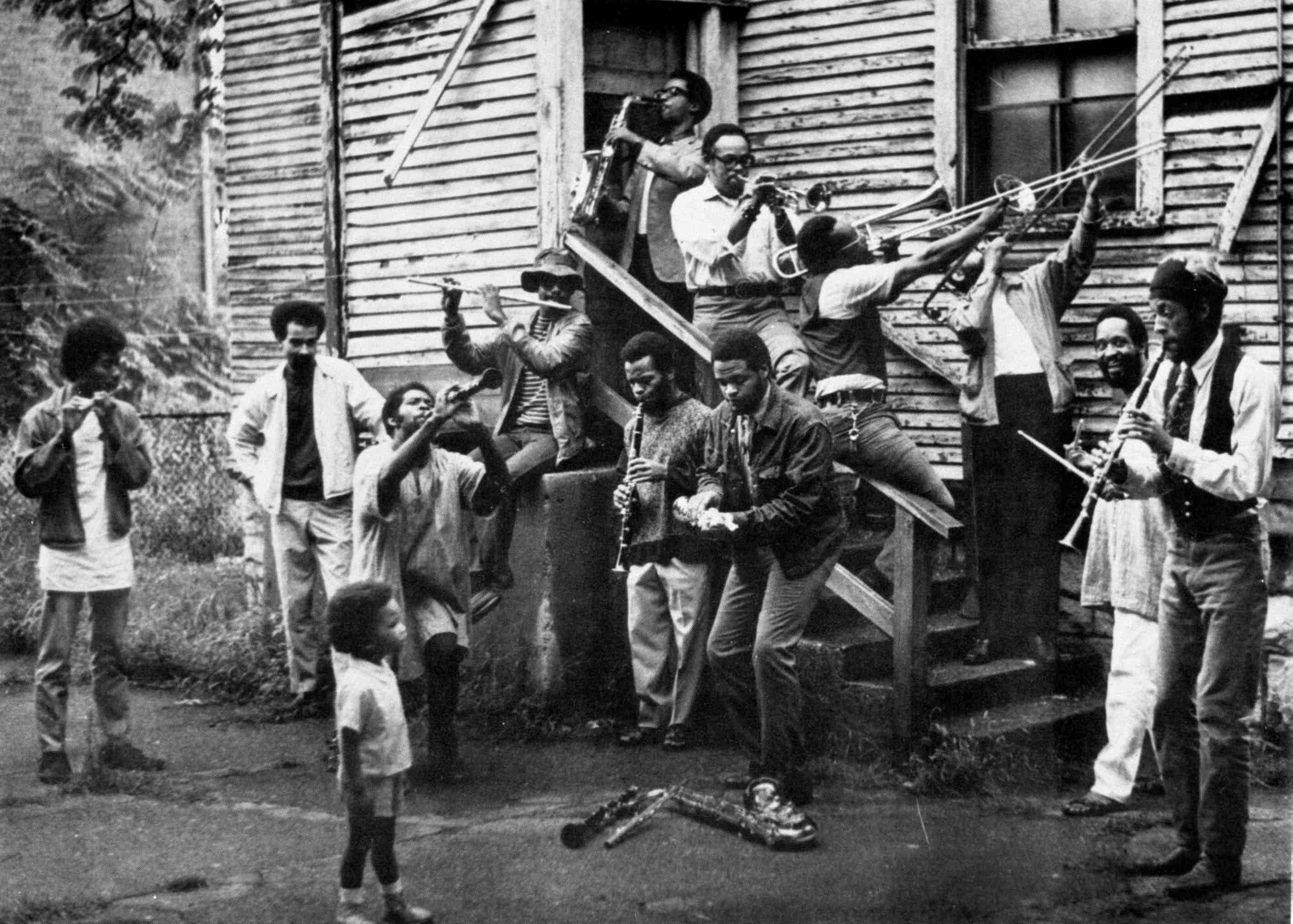 A black and white photograph of a crowd of men joyously playing all kinds of musical instruments on a porch while a young boy looks on