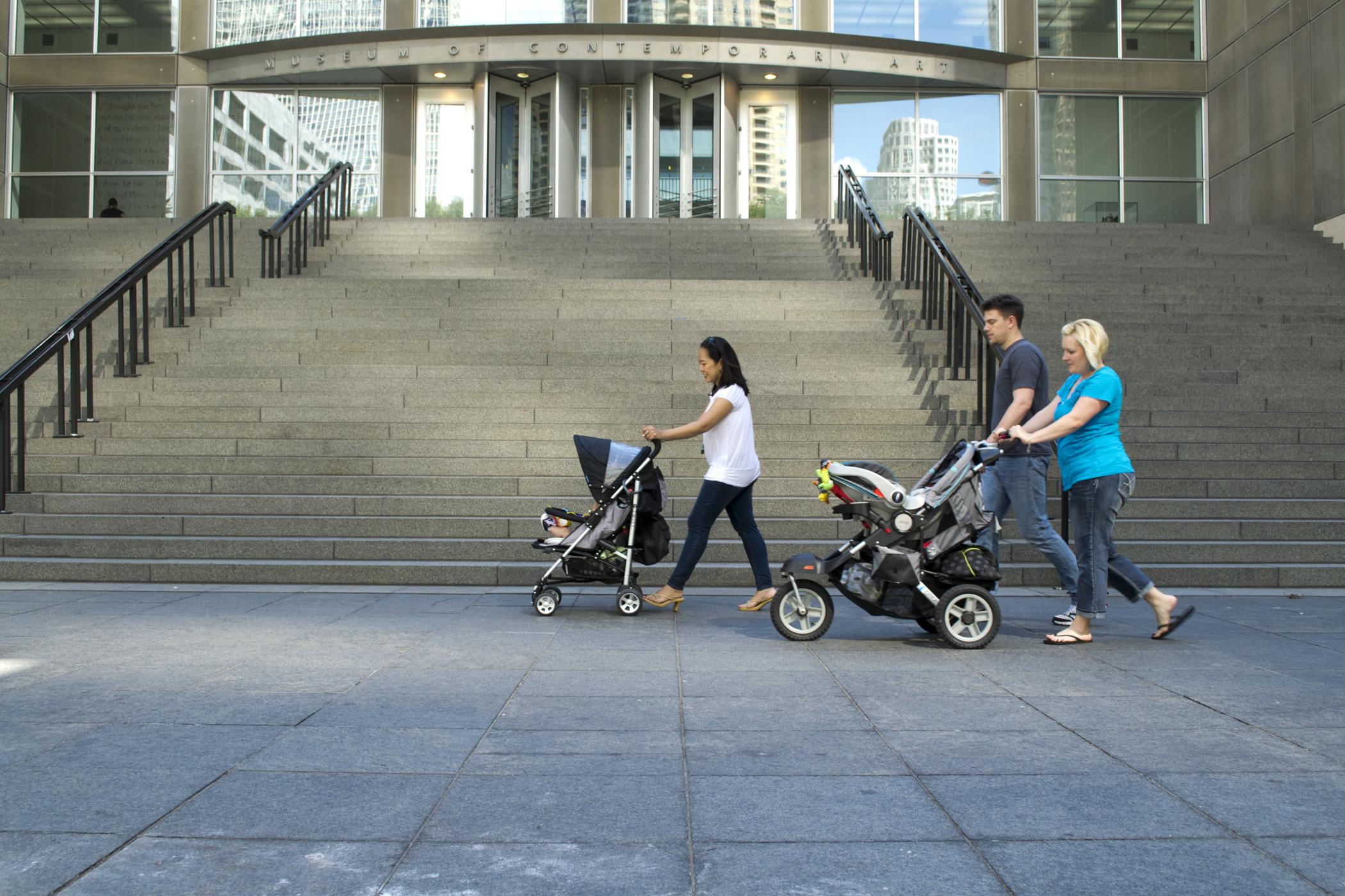 Three adults walk across the MCA plaza on a sunny day, two are pushing strollers.