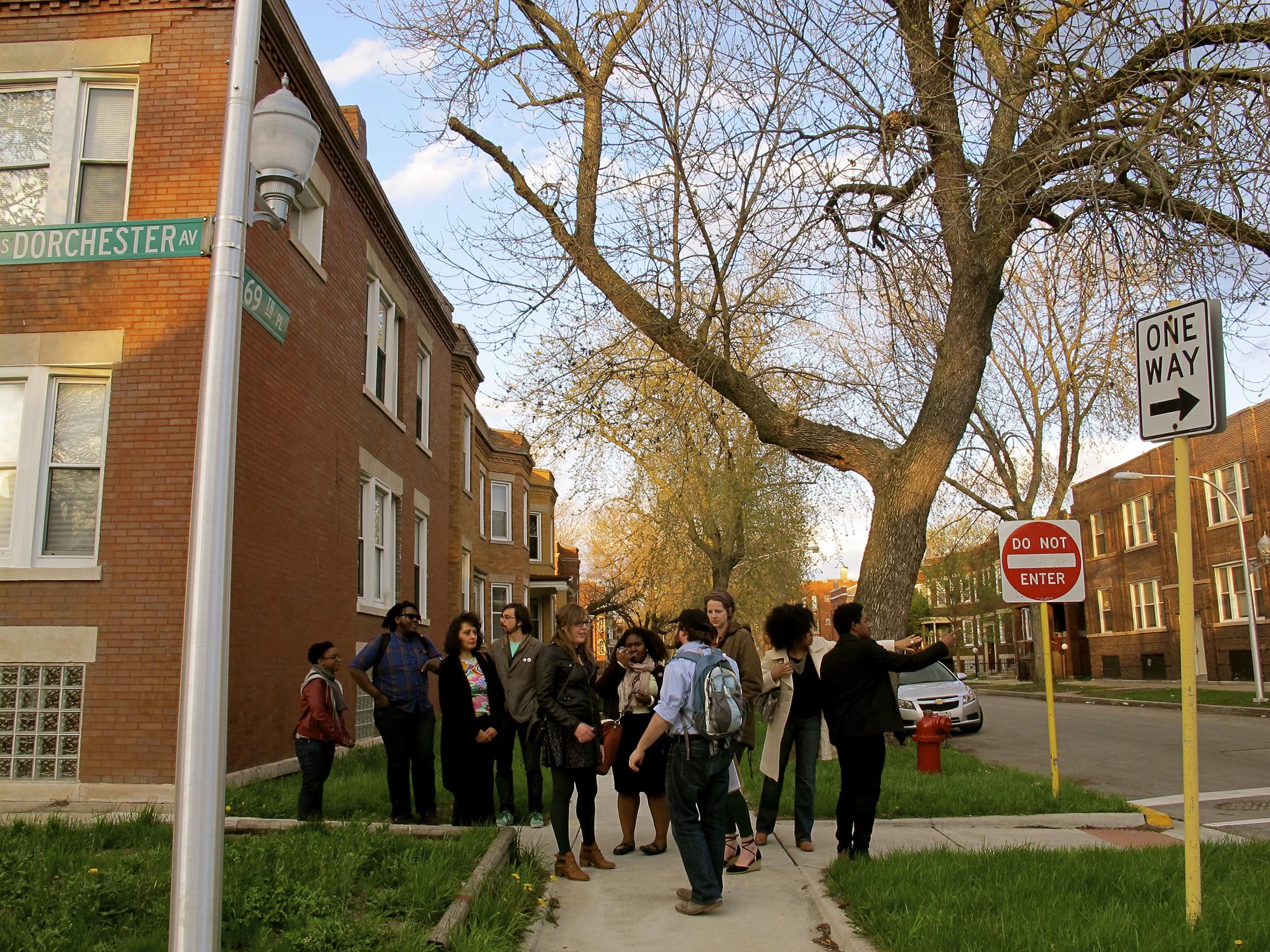 A group of young people of different races are clustered at the end of a sidewalk as a man wearing a backpack leads them; the street signs in the image say "Dorchester Avenue" and "69th Place."