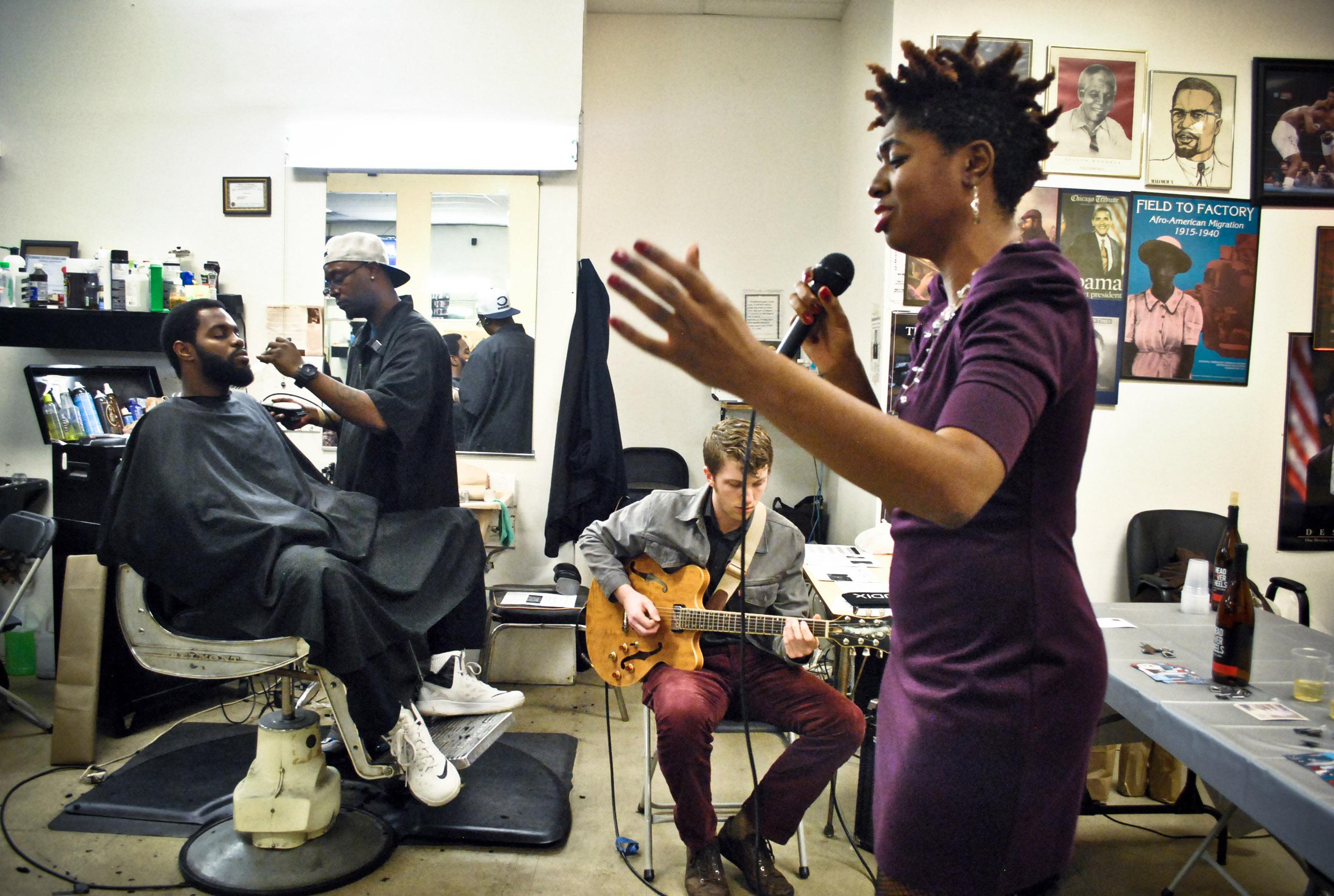 A young black woman sings into a microphone in a barbershop; a young white man sits behind her playing electric guitar. To his left a black man sits in a barber's chair getting his hair cut.