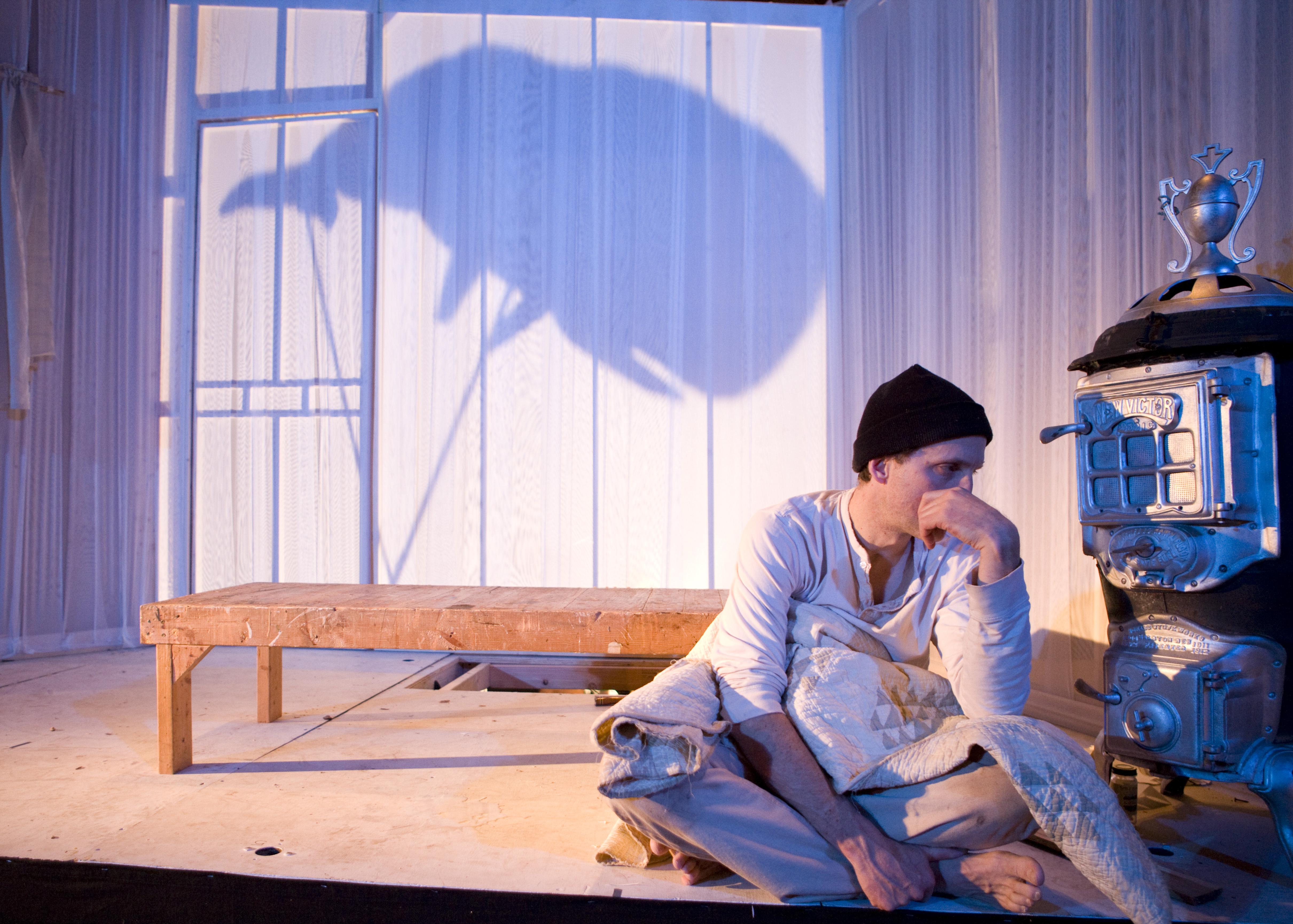 A man sits dejectedly on a small stage in front of a wooden bench and a wood-burning stove. A whale shadow puppet appears on the white curtain backdrop.