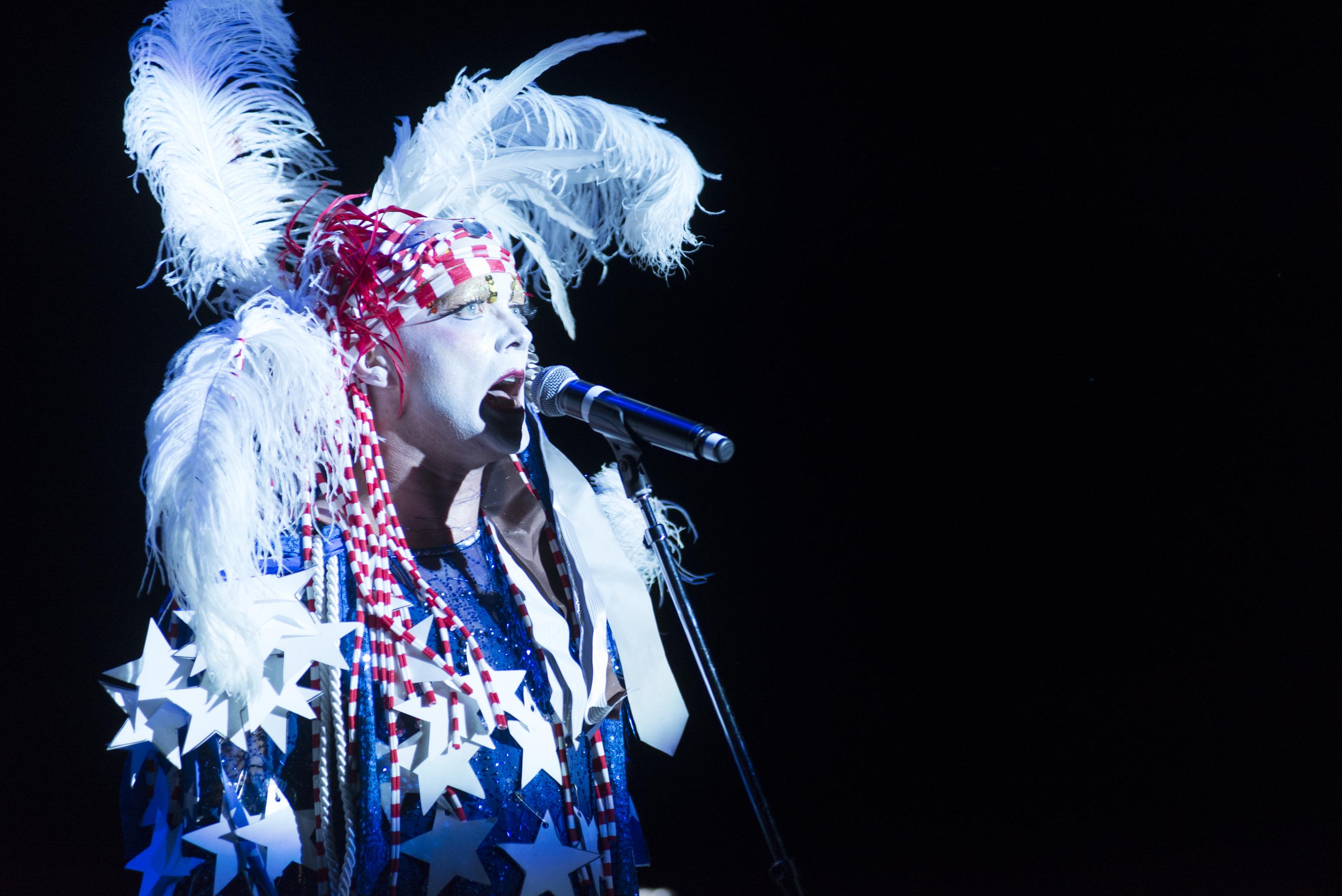 A person sings at a microphone on a pitch-black stage, wearing a white feather headdress, a red-striped bandana, and blue dress strung with white paper stars.