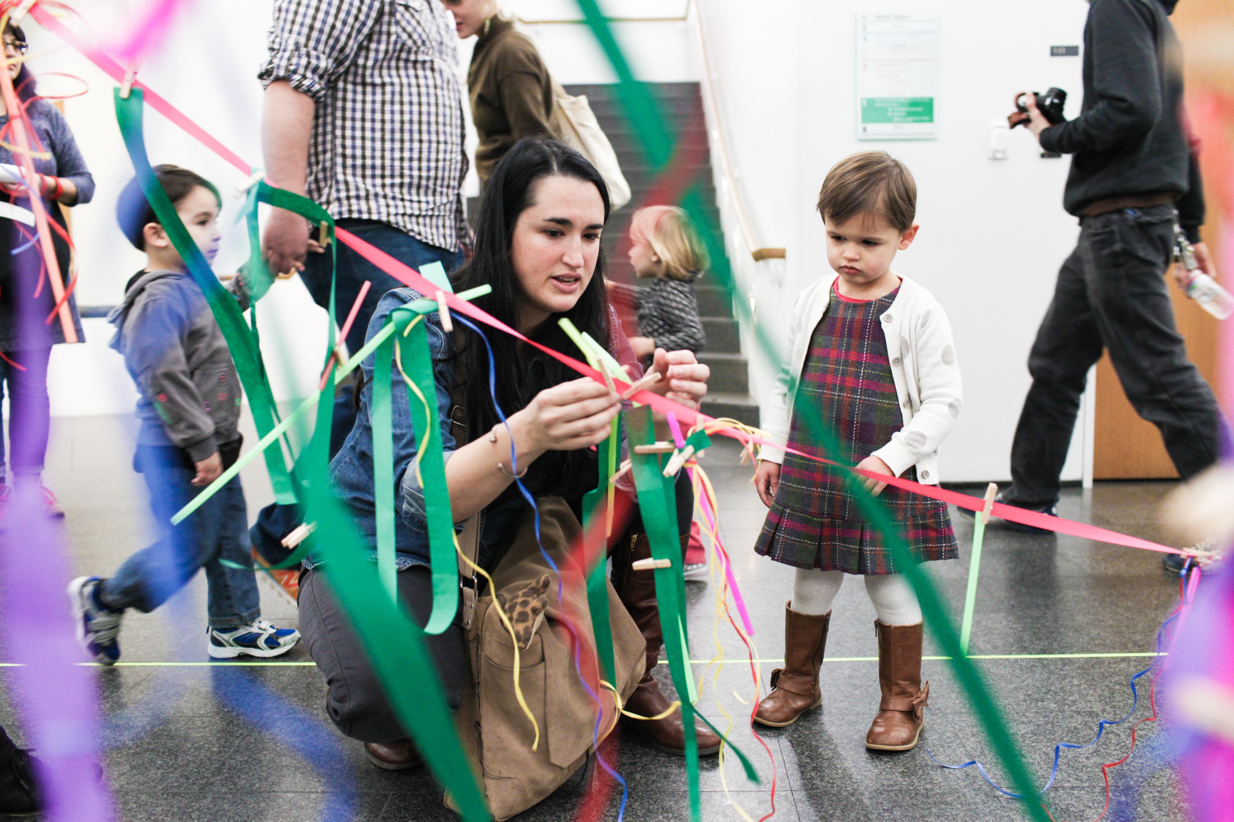 A woman ties a knot of ribbon onto another while a young child watches.