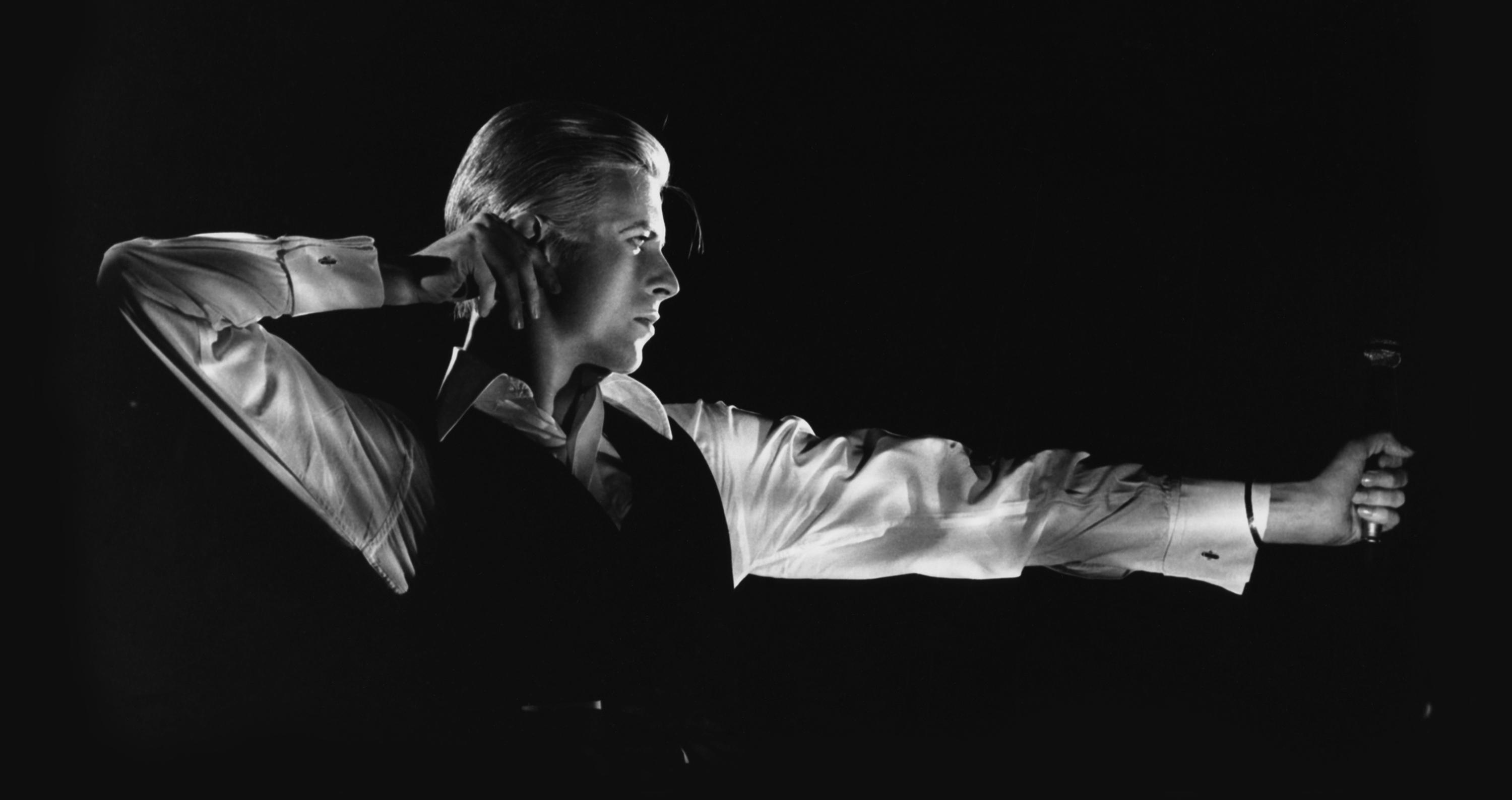 Black-and-white portrait of David Bowie in profile, arms positioned as though shooting a bow and arrow, a microphone in his extended arm