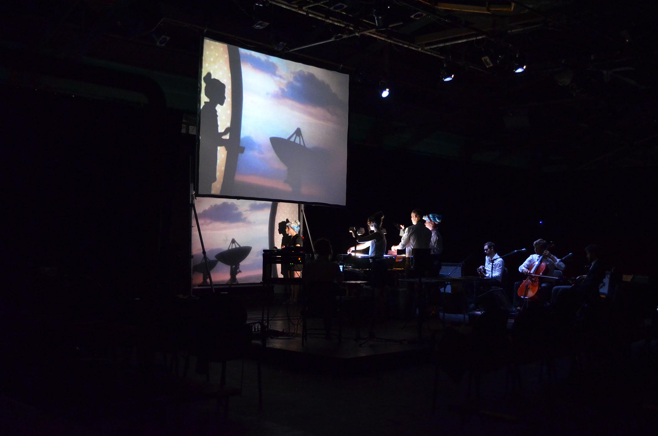 On a dark stage, technicians use overhead projectors to cast ta woman's silhouette onto an image of a sunset that appears in the background.