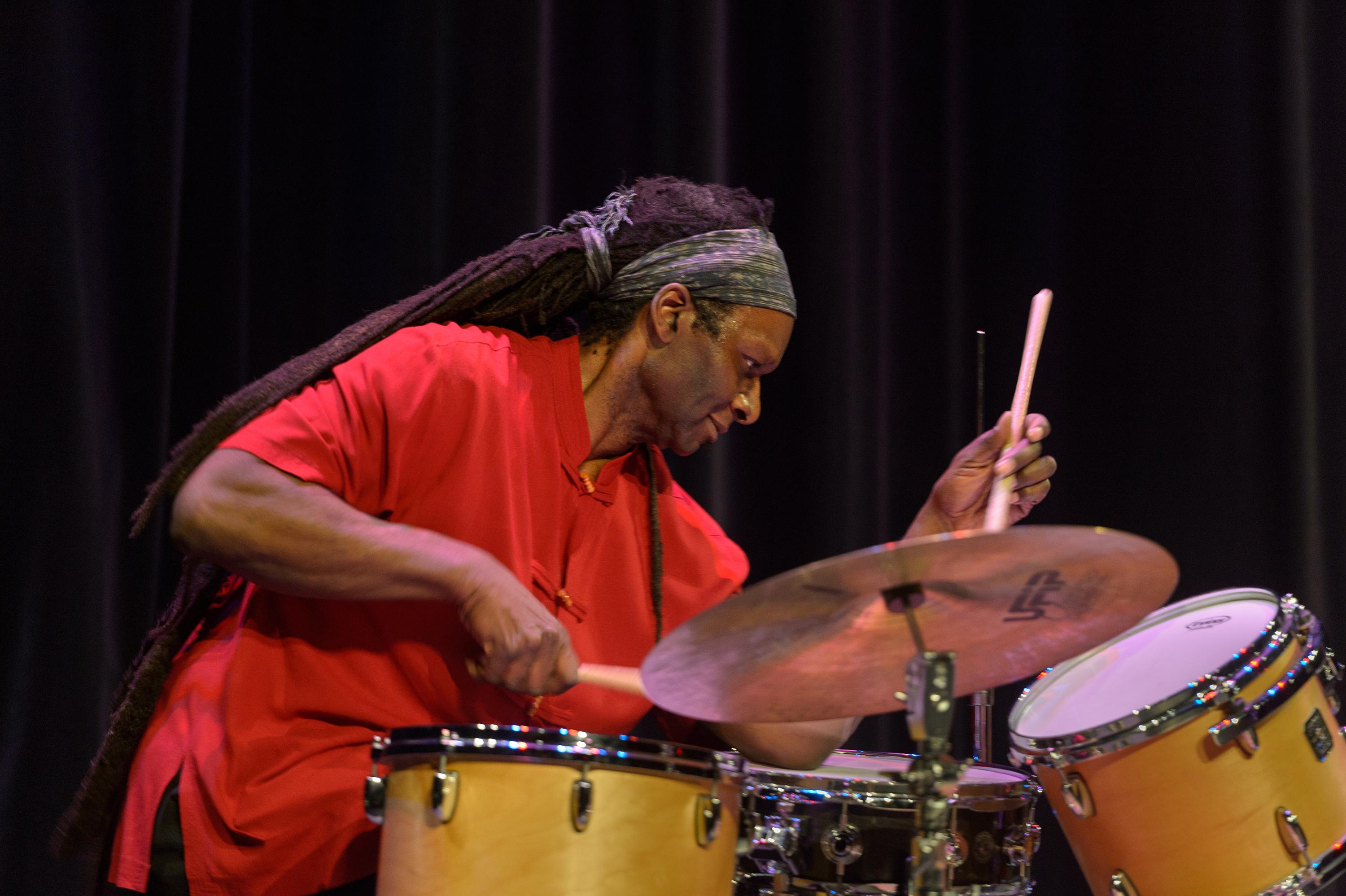 A man with dreadlocks that hang past his waist playing a drum kit in front of a black curtain