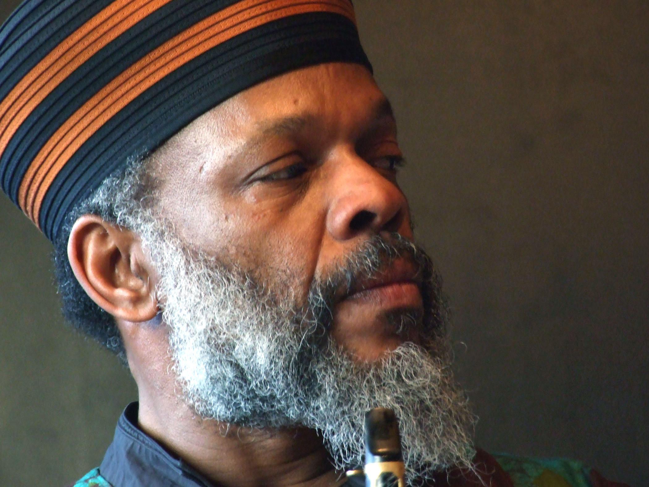 Portrait of a man with a long grey beard wearing a black and orange striped hat, the tip of a woodwind reed near his chin