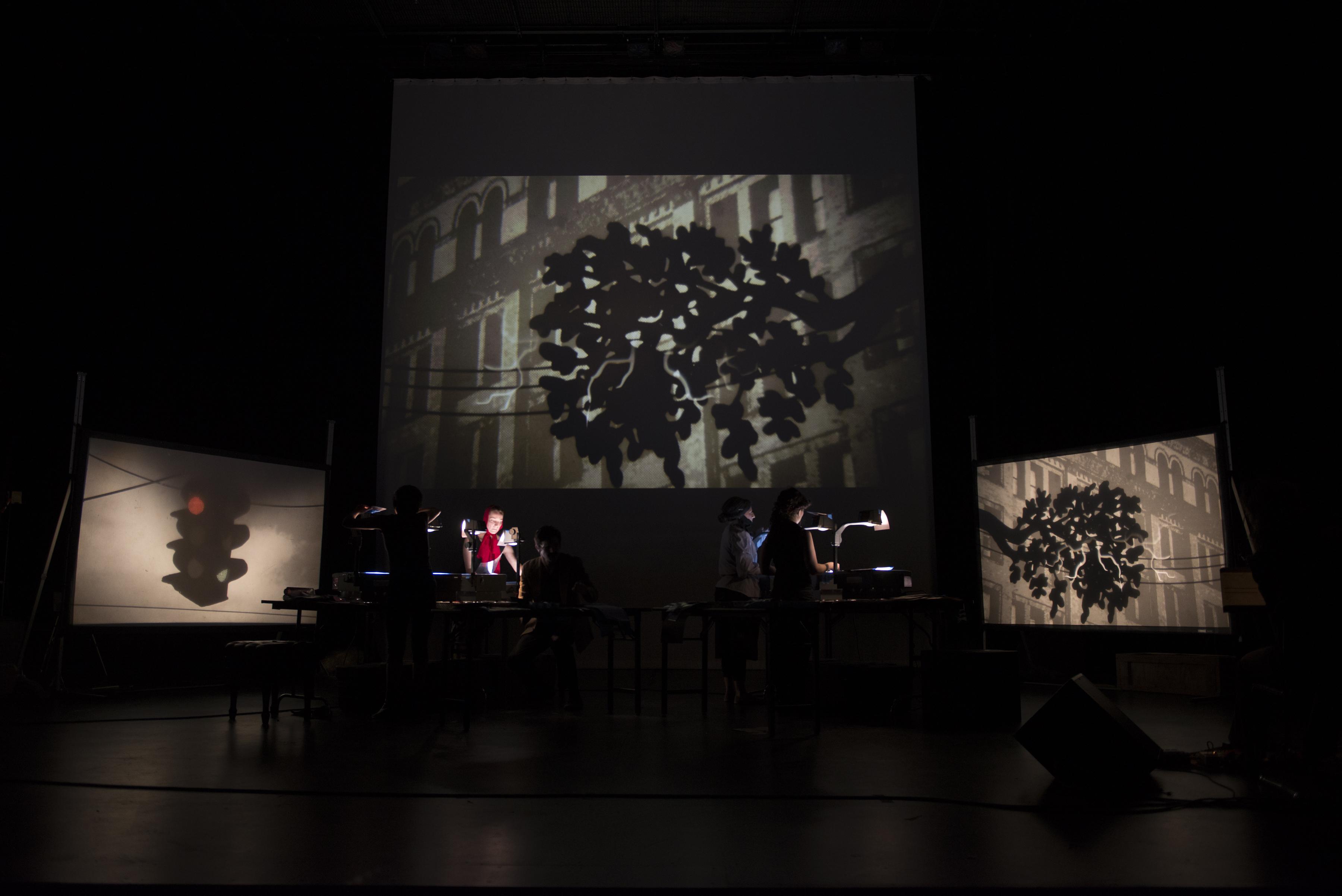 Technicians use overhead projectors to cast a street scene onto three large screens in the background.
