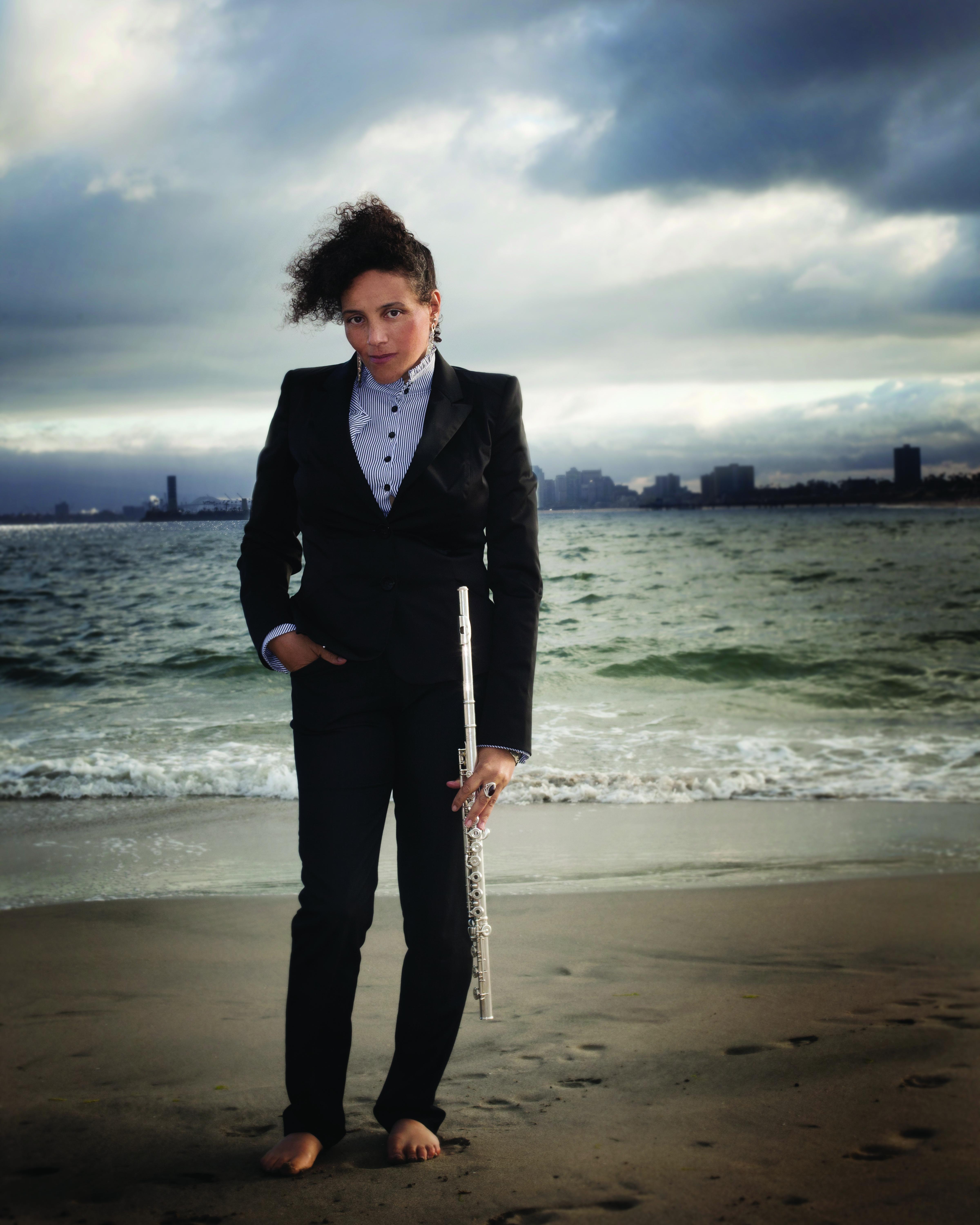 A person in a black suit stands barefoot on a beach while holding a flute.