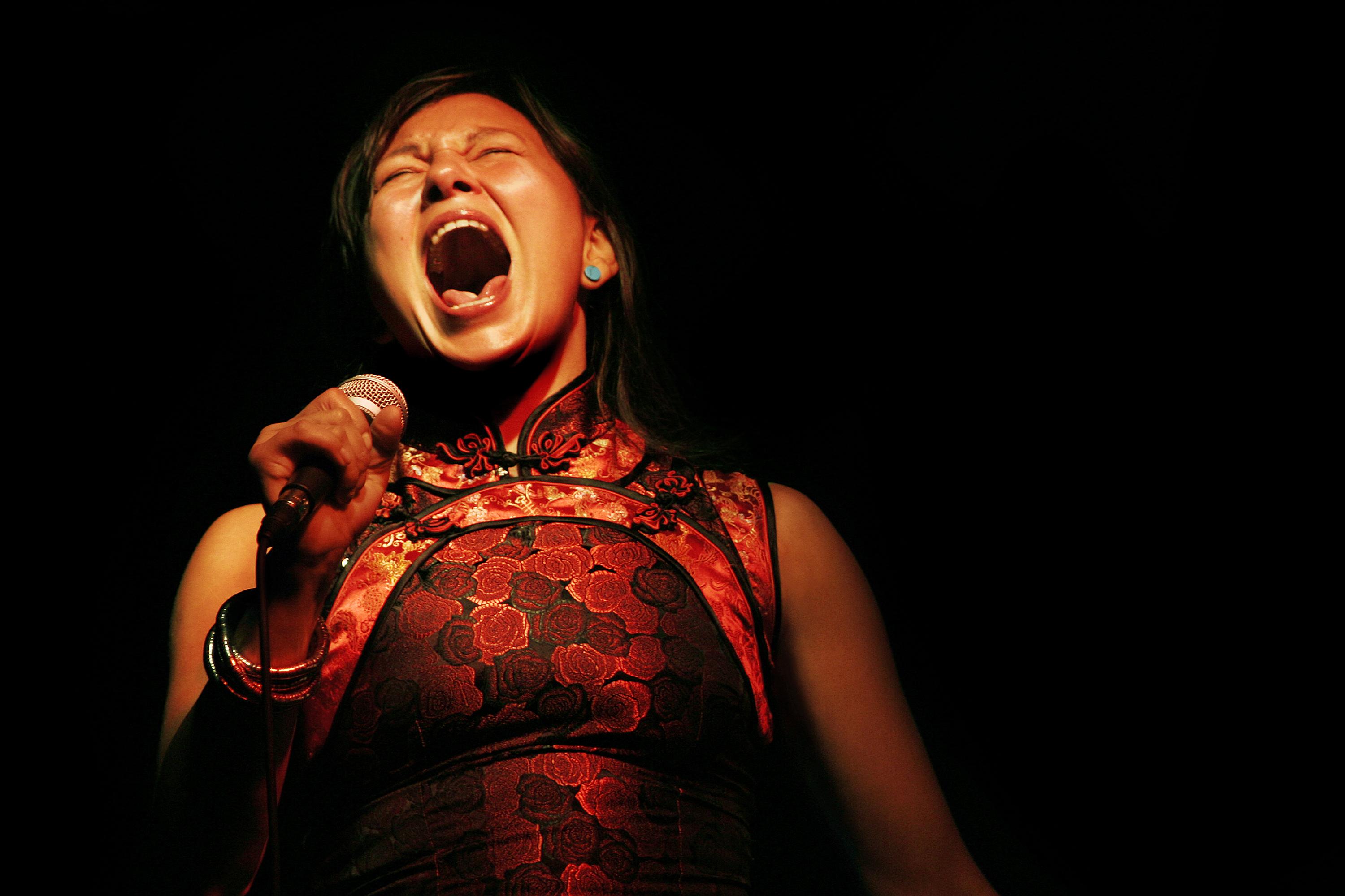 A woman in in Asian clothing sings into her microphone on a pitch-black stage