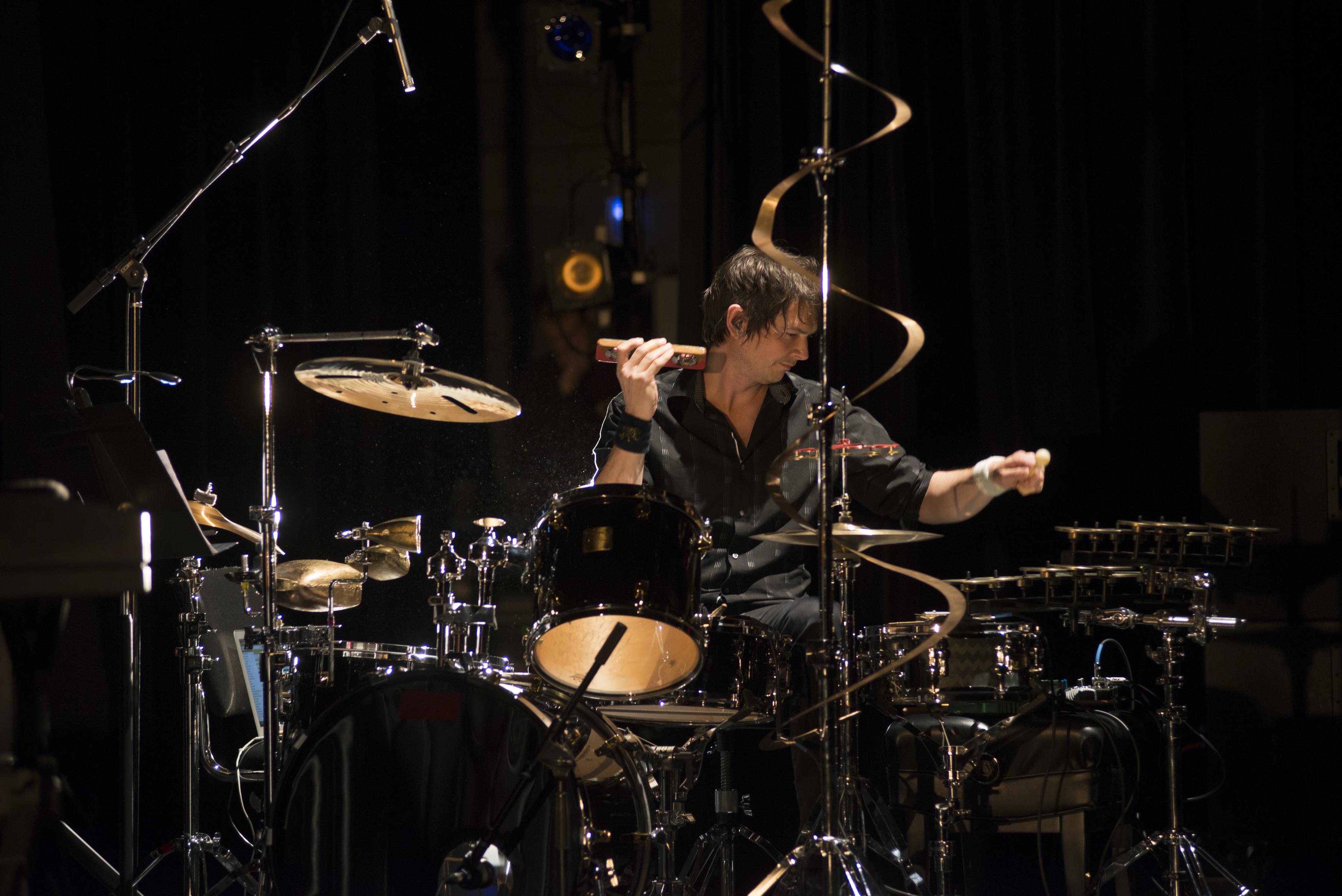 A man sits at a drum set and unusual percussion instruments. He holds a wood block to his ear.