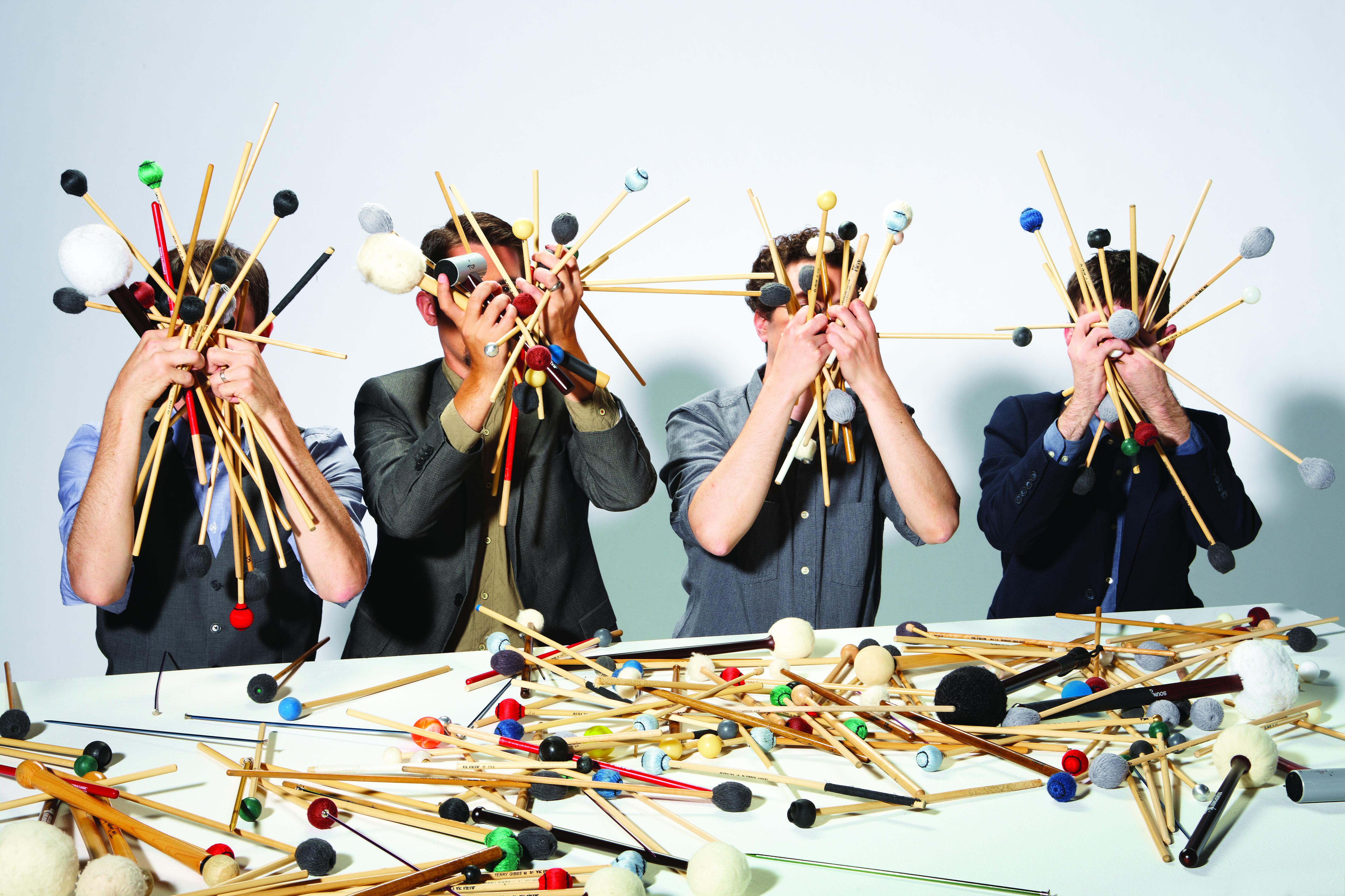 Four men in dress shirts and suit jackets hold handfuls of drumsticks and mallets in front of their faces while sitting at a table also covered with drumsticks and mallets.