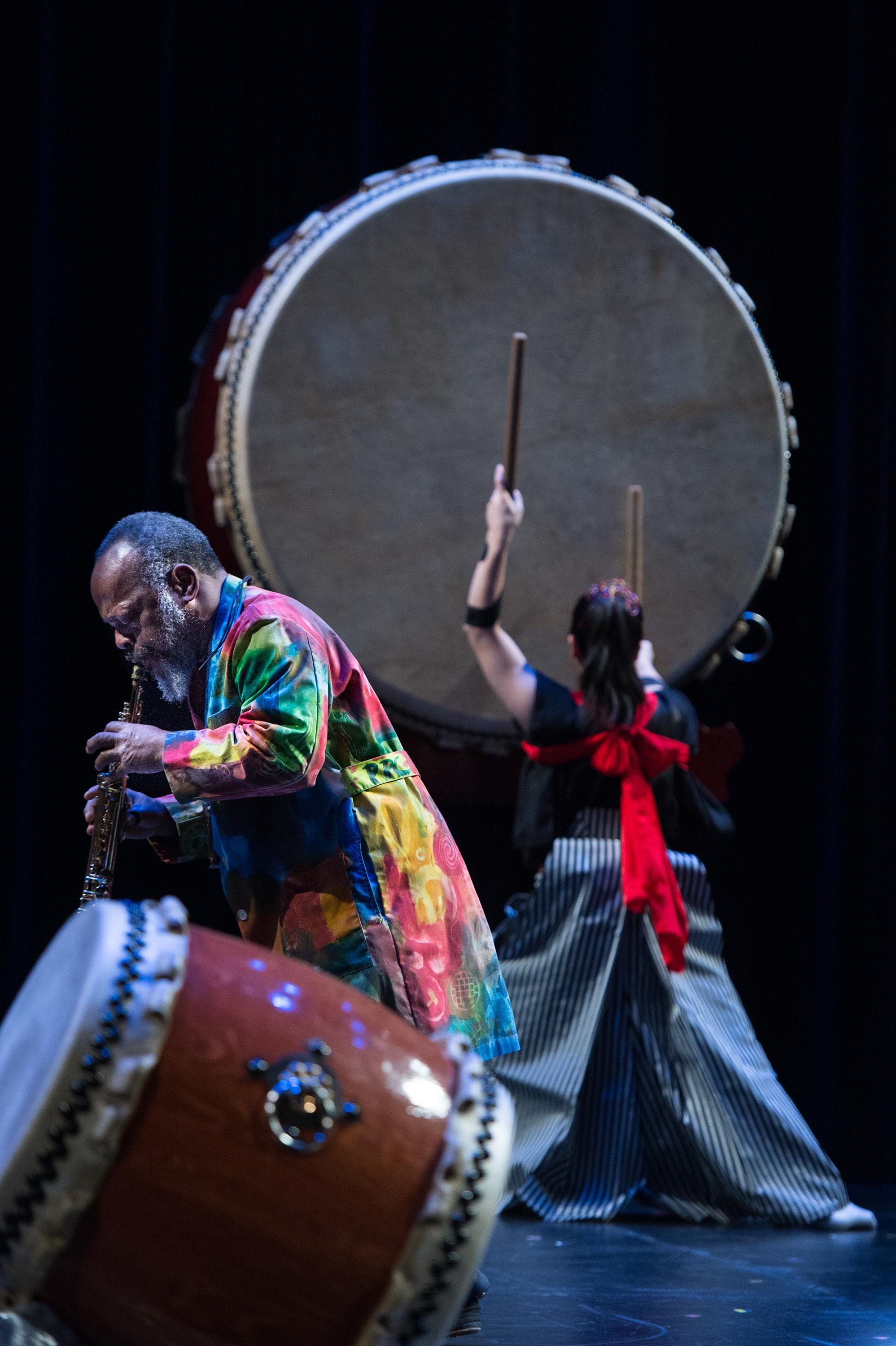 A man plays a soprano saxophone on stage while a woman beats an enormous taiko drum in the background.