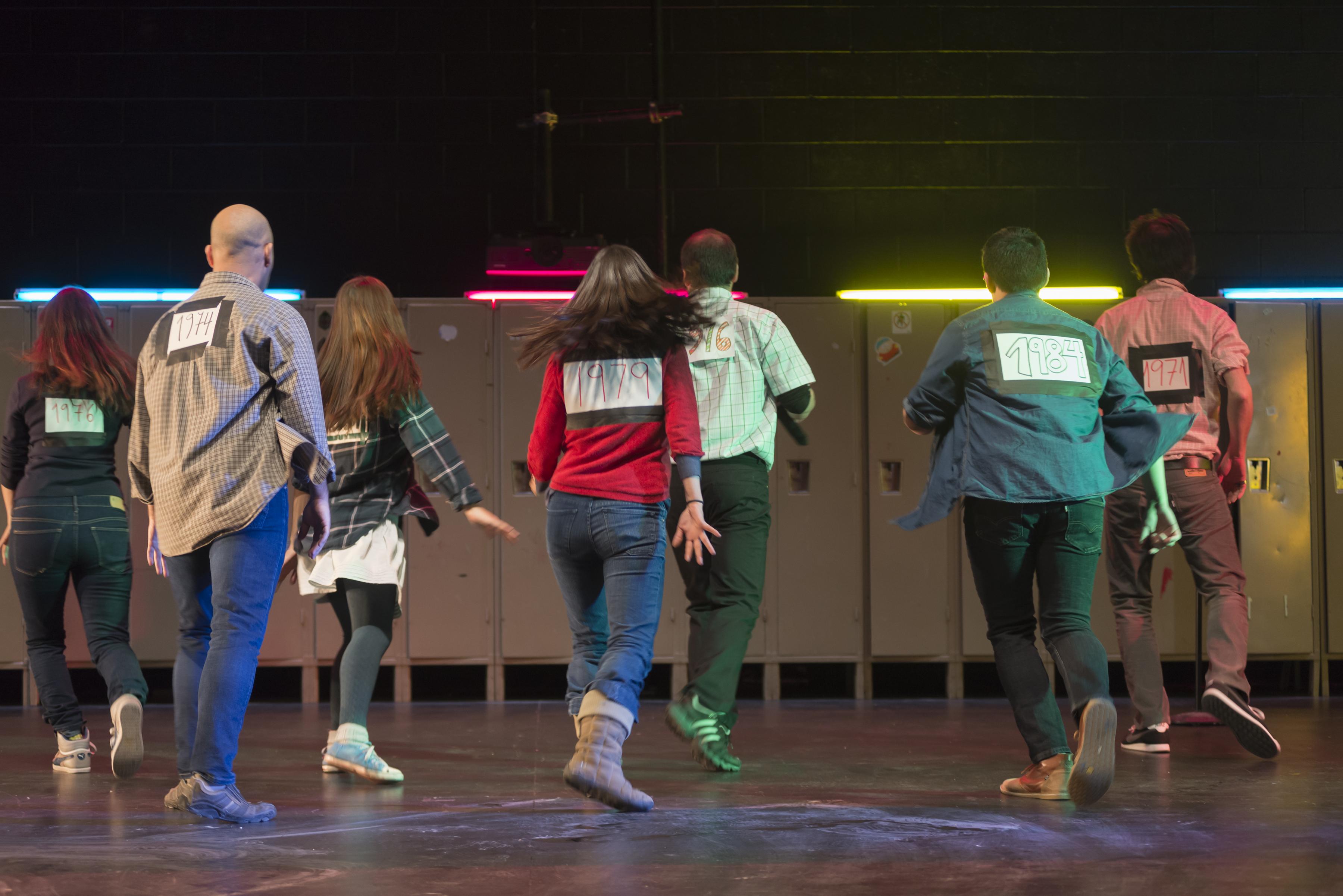 A performance still shows seven actors walking away from the audience toward lockers at the back of the stage.