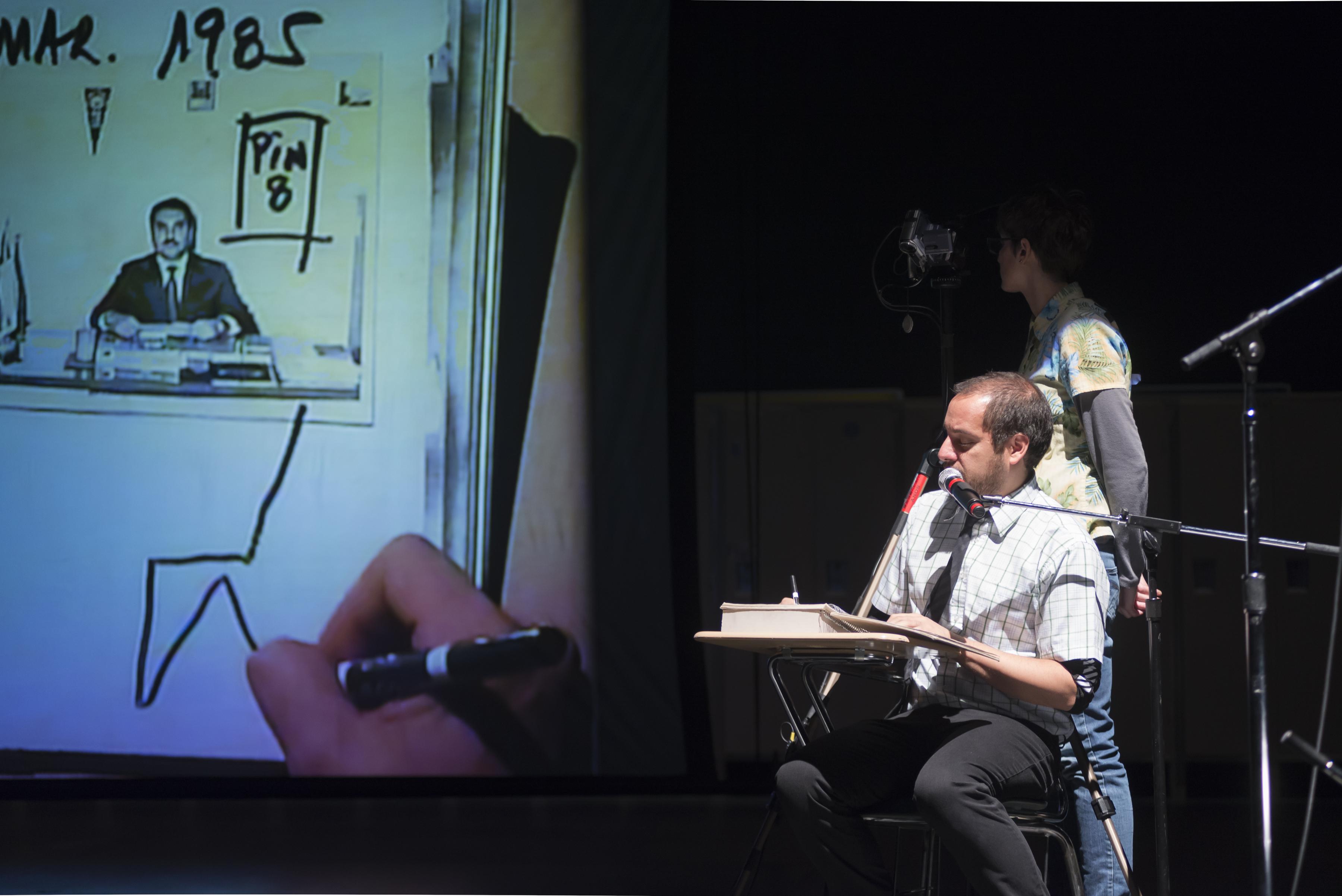 A performance still shows one actor watching a projection of what a seated actor draws at his desk.