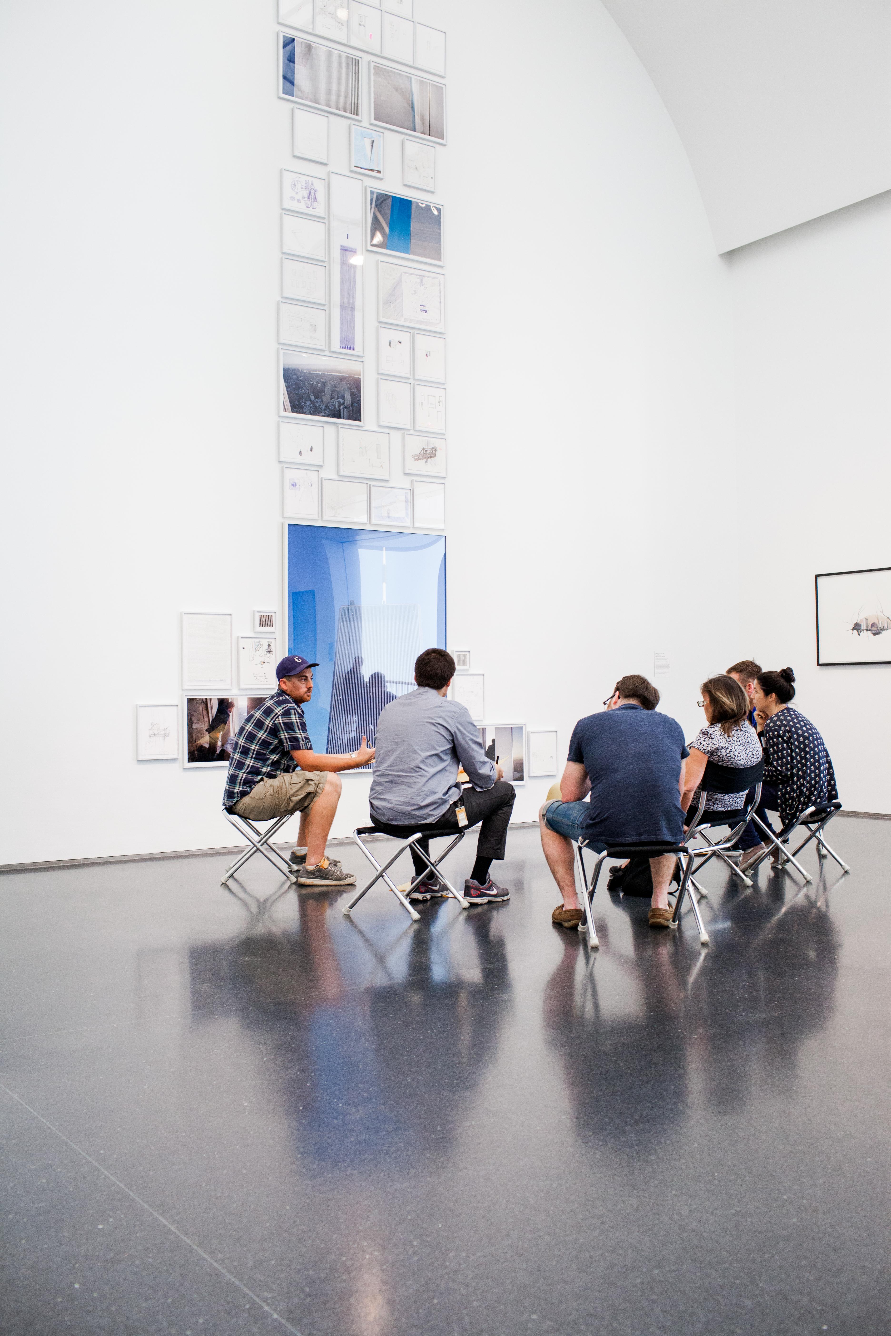 Six teachers sit on folding stools in front of an installation of drawings and photographs.