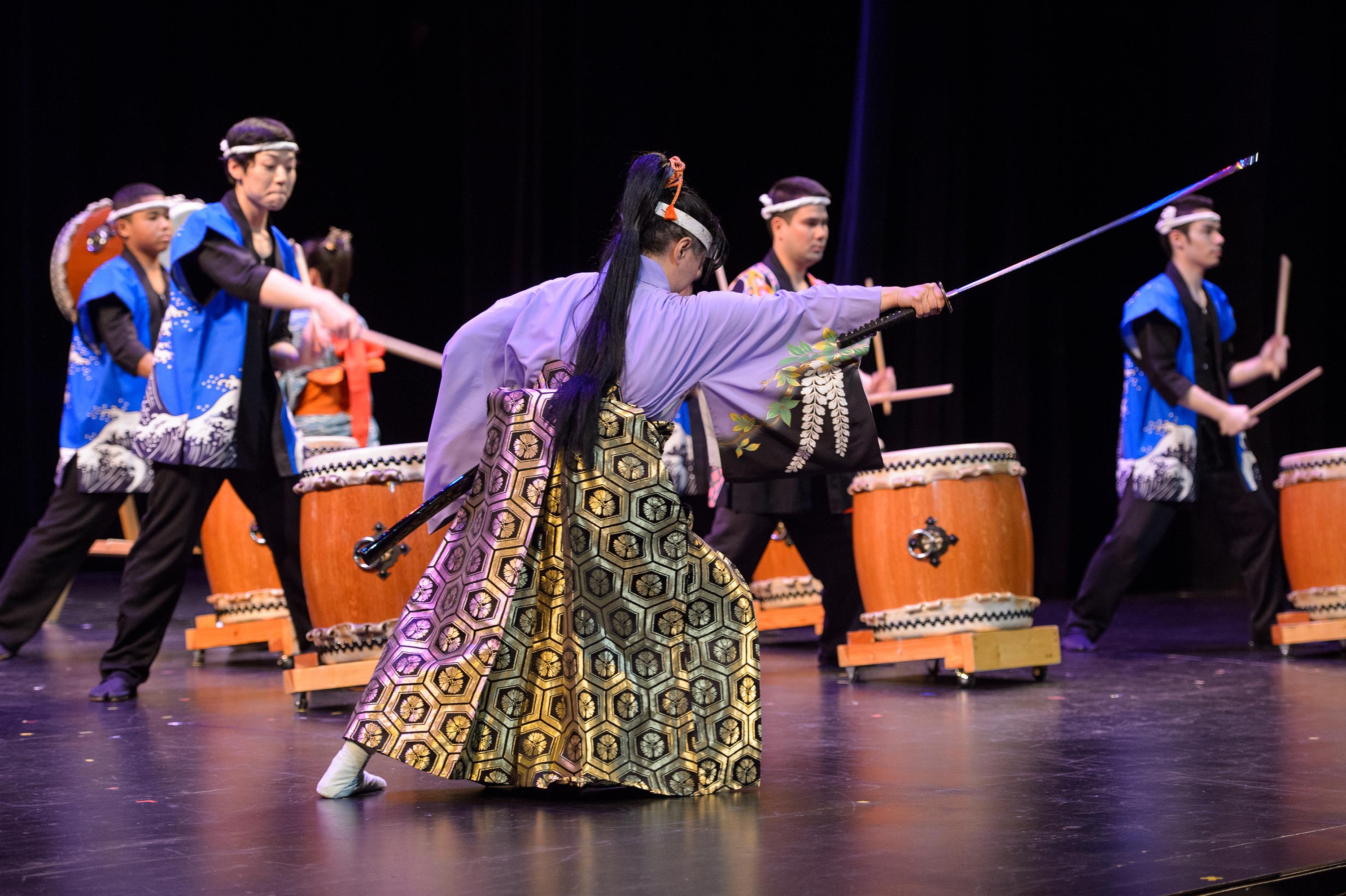 On stage, a woman in traditional Japanese clothing lunges with a long-handled sword while young male drummers play *taiko* drums behind her.