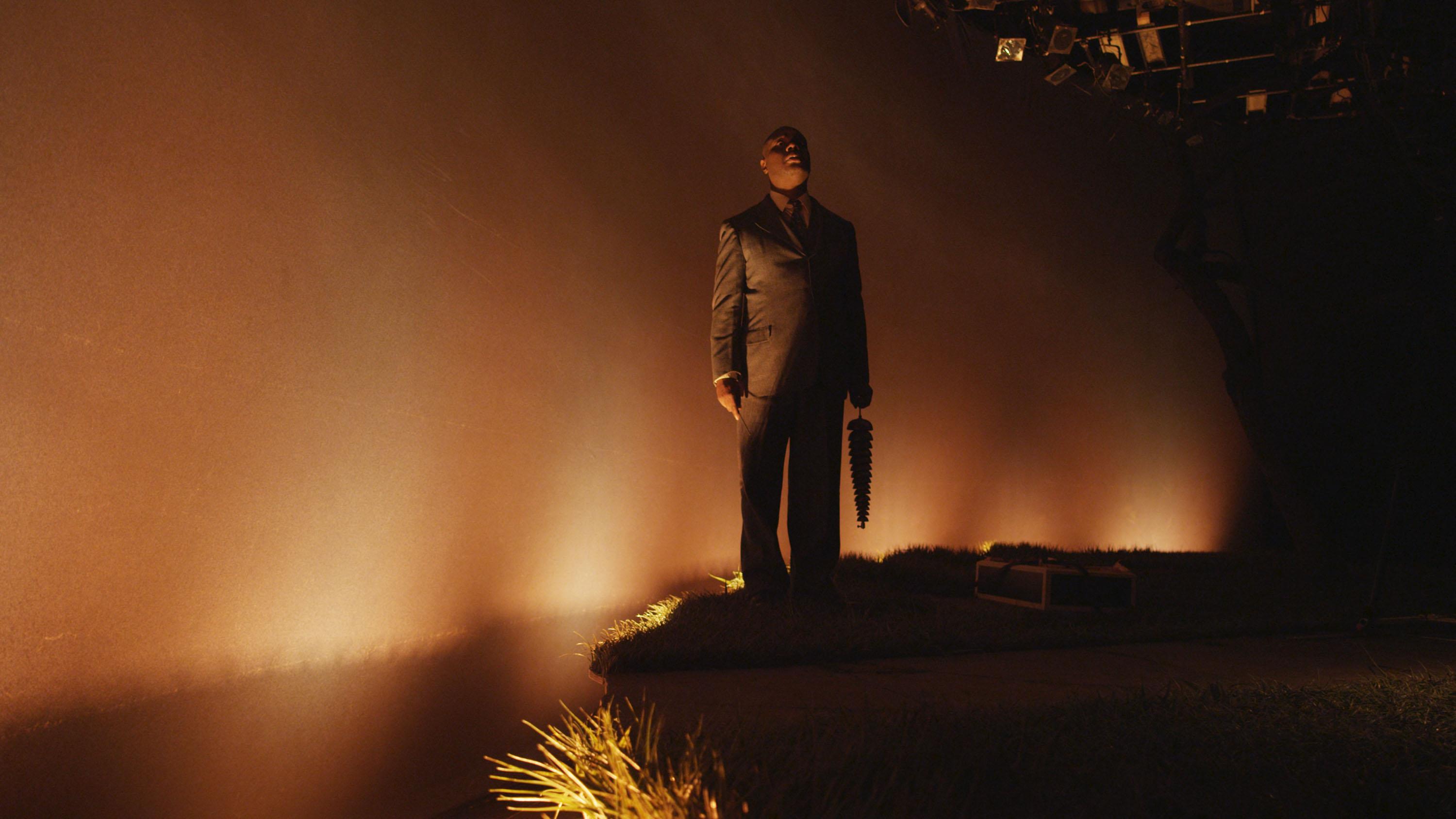 Video still shows a man wearing a suit on an eerie, dimly lit stage.