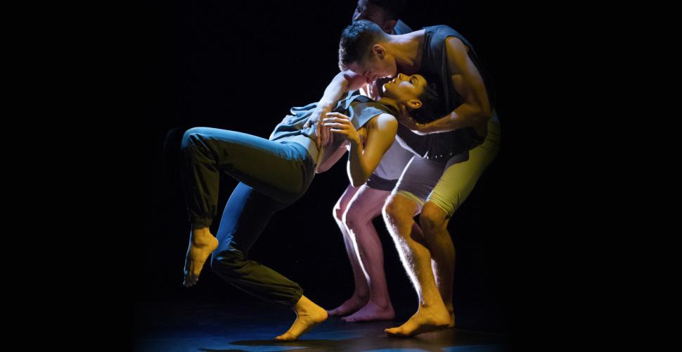 A female dancer dressed in sweats leans back toward a male dancer, who holds her upright with a hand behind her neck. A third dancer stands behind, their face and torso blocked from view.