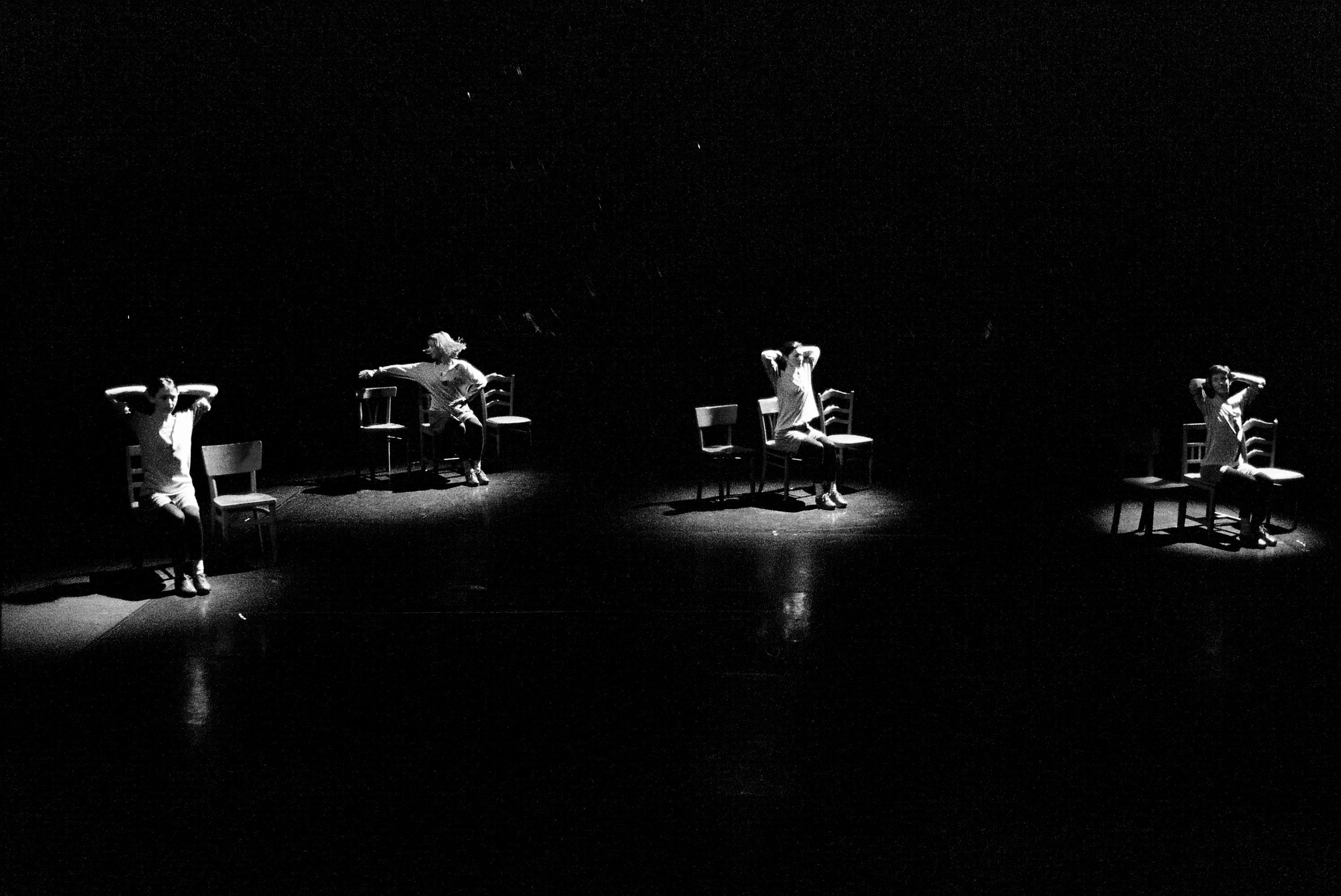 Black-and-white performance still of four women on a pitch-black stage, each one illuminated by a spotlight and dancing in a chair