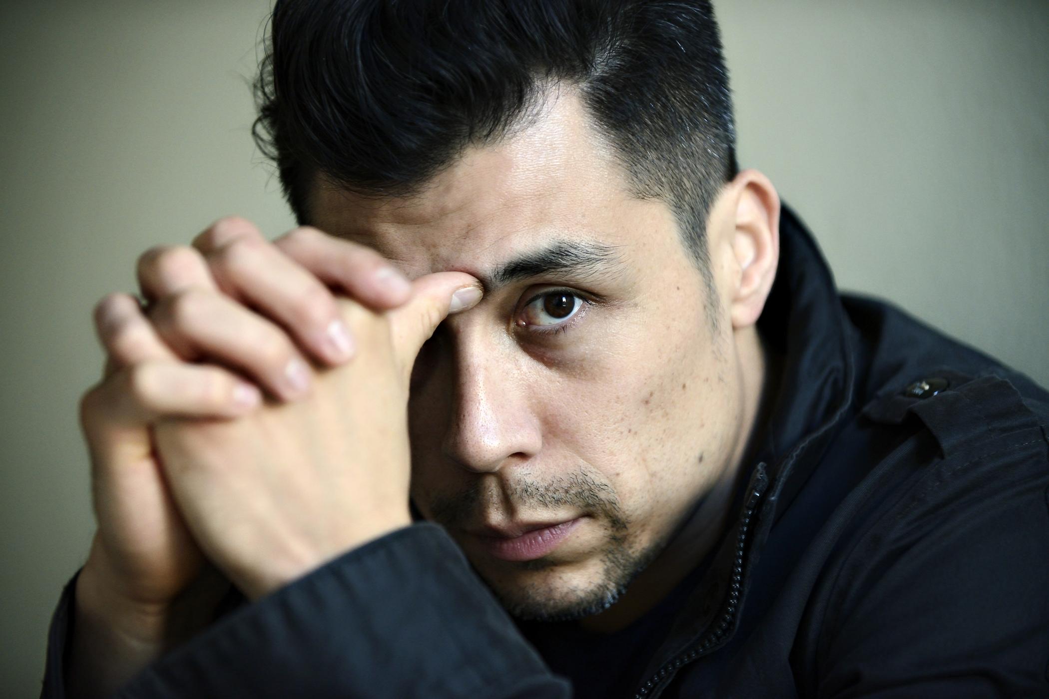 A light-skinned man poses with hands folded, thumb pressed against the bridge of his nose. He looks out at the viewer.