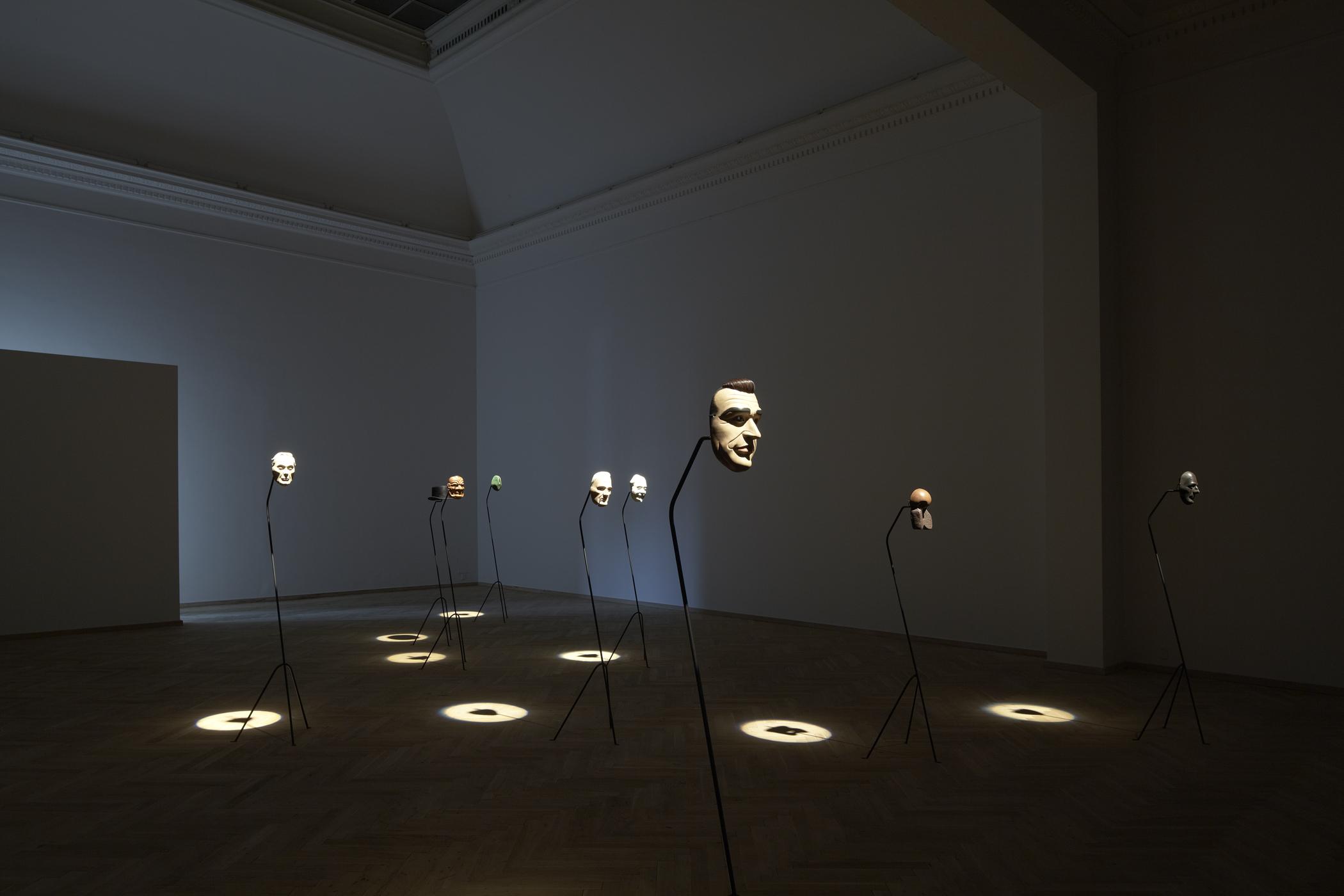Installation view of a dark room with nine human-face masks illuminated by spotlights