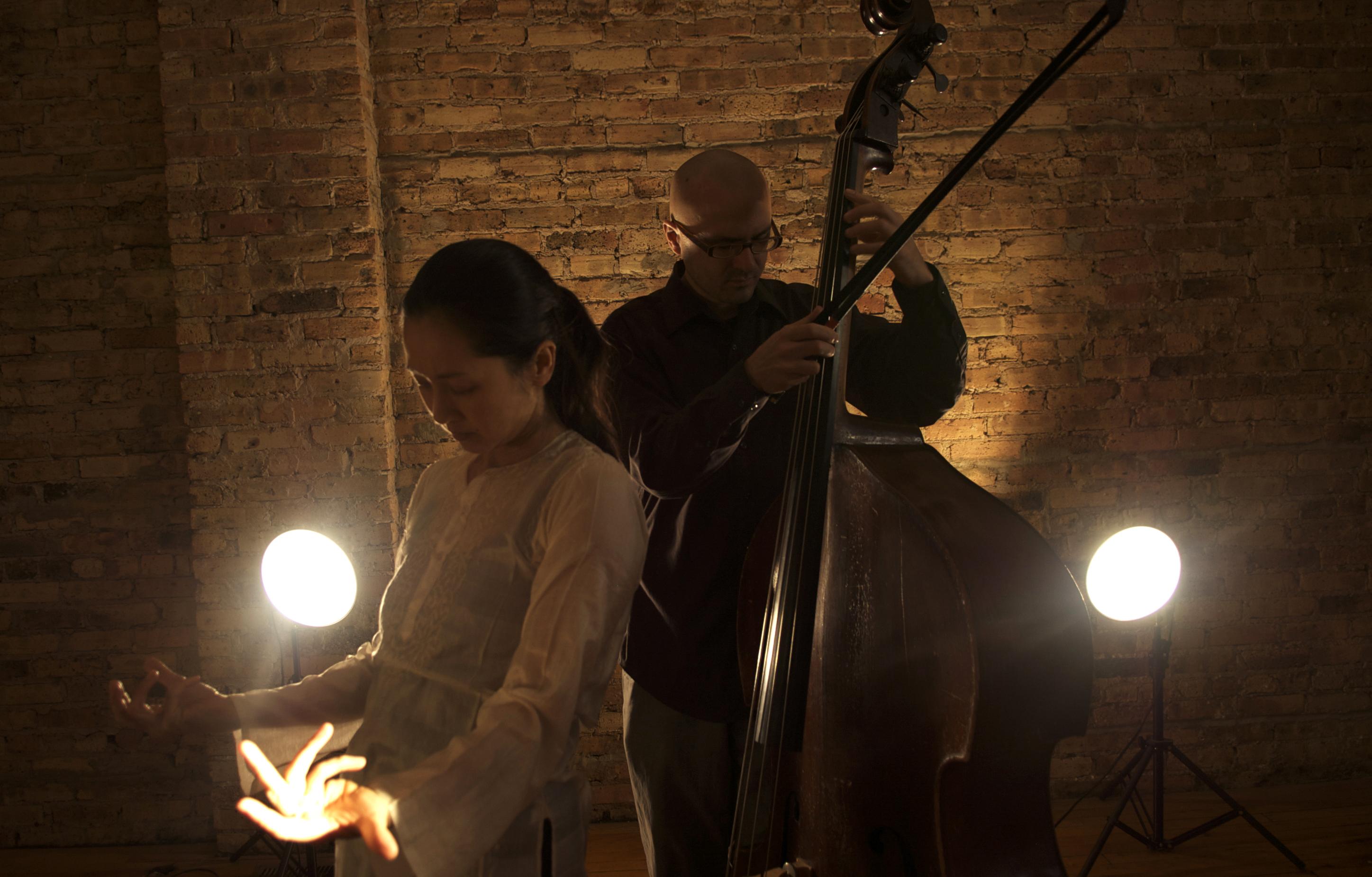 A woman and man in a dark room are backlit by two spotlights. He plays a string bass while she gestures with both arms, empty handed and fingers curled.