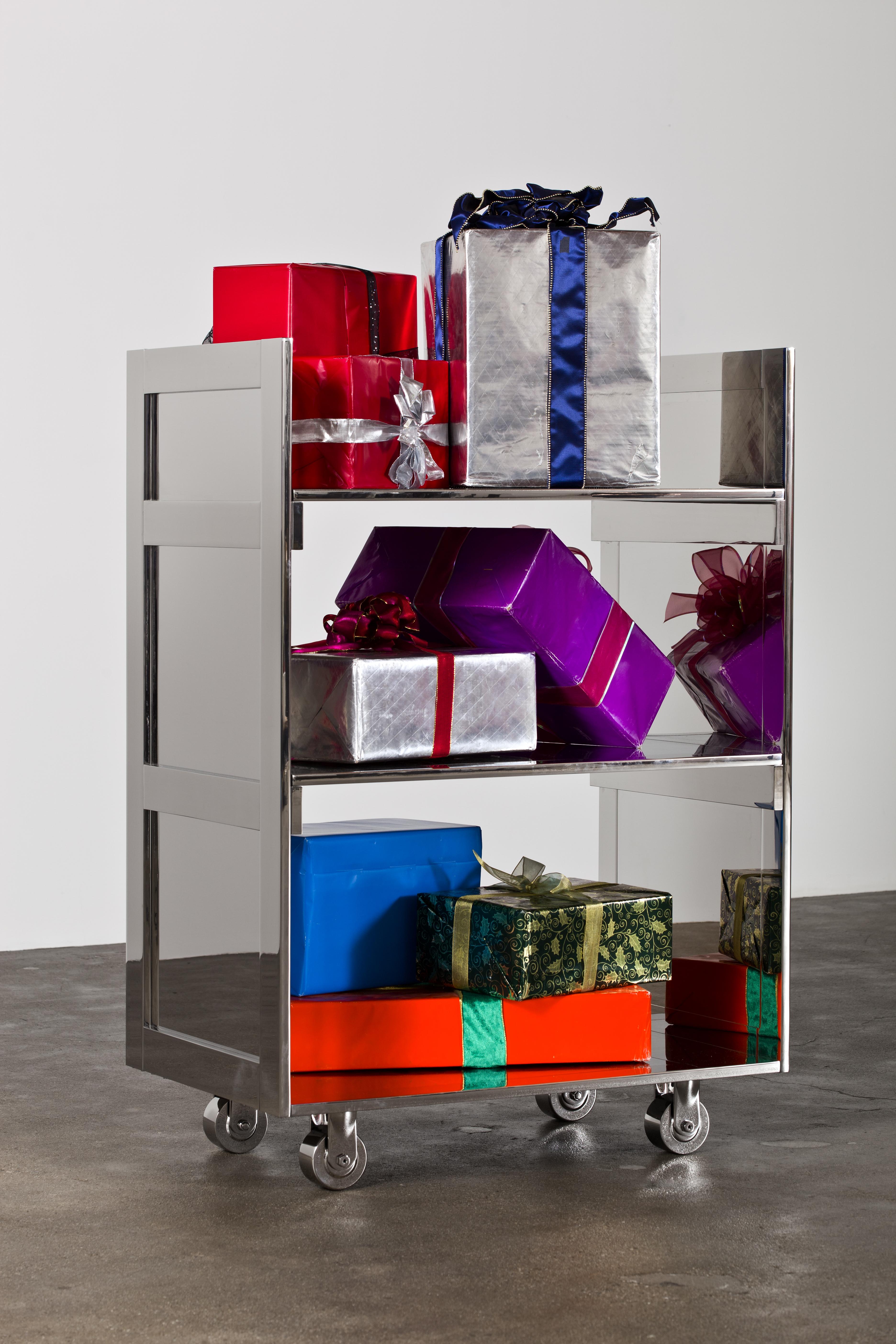 A chrome metal cart with three shelves holds boxes gift wrapped in colorful, shiny paper and ribbons with bows.