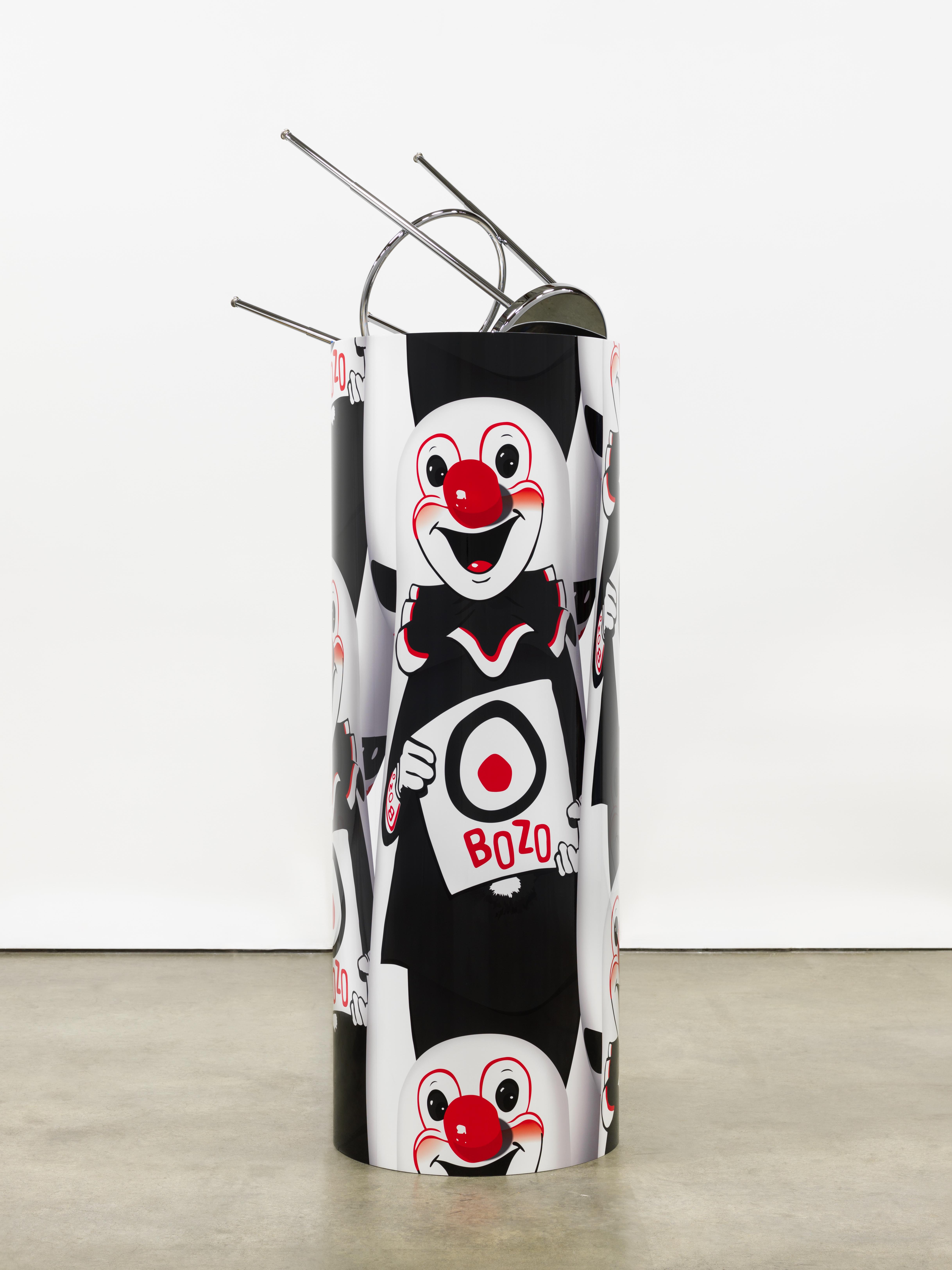 A cylinder is wrapped with the repeating image of a white, red, and black clown holding a target with "BOZO" written below. An upside-down stool protrudes from the top of the sculpture.