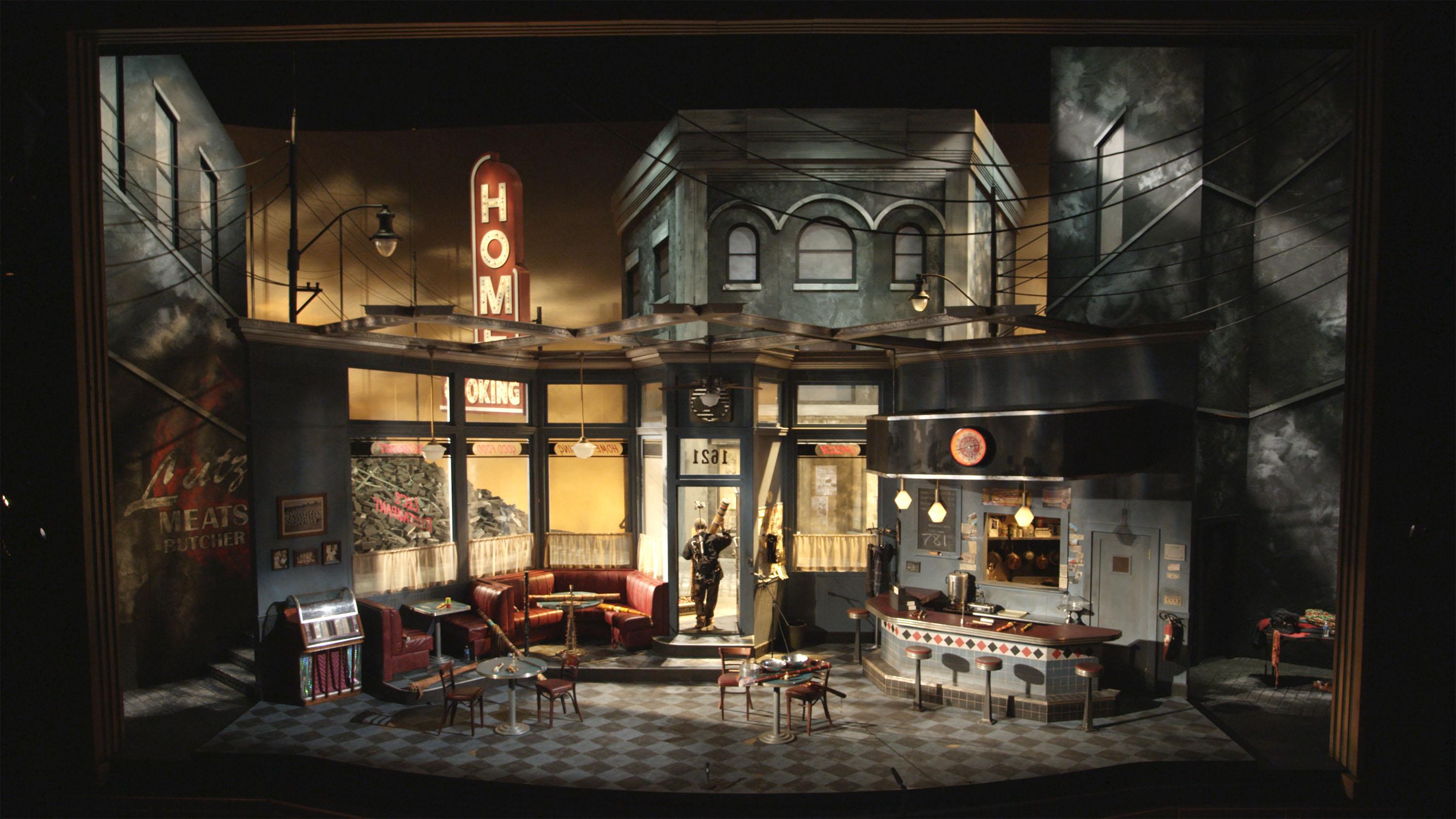 A shadowy stage set featuring a 1950s diner amid row houses, with a single spotlit performer