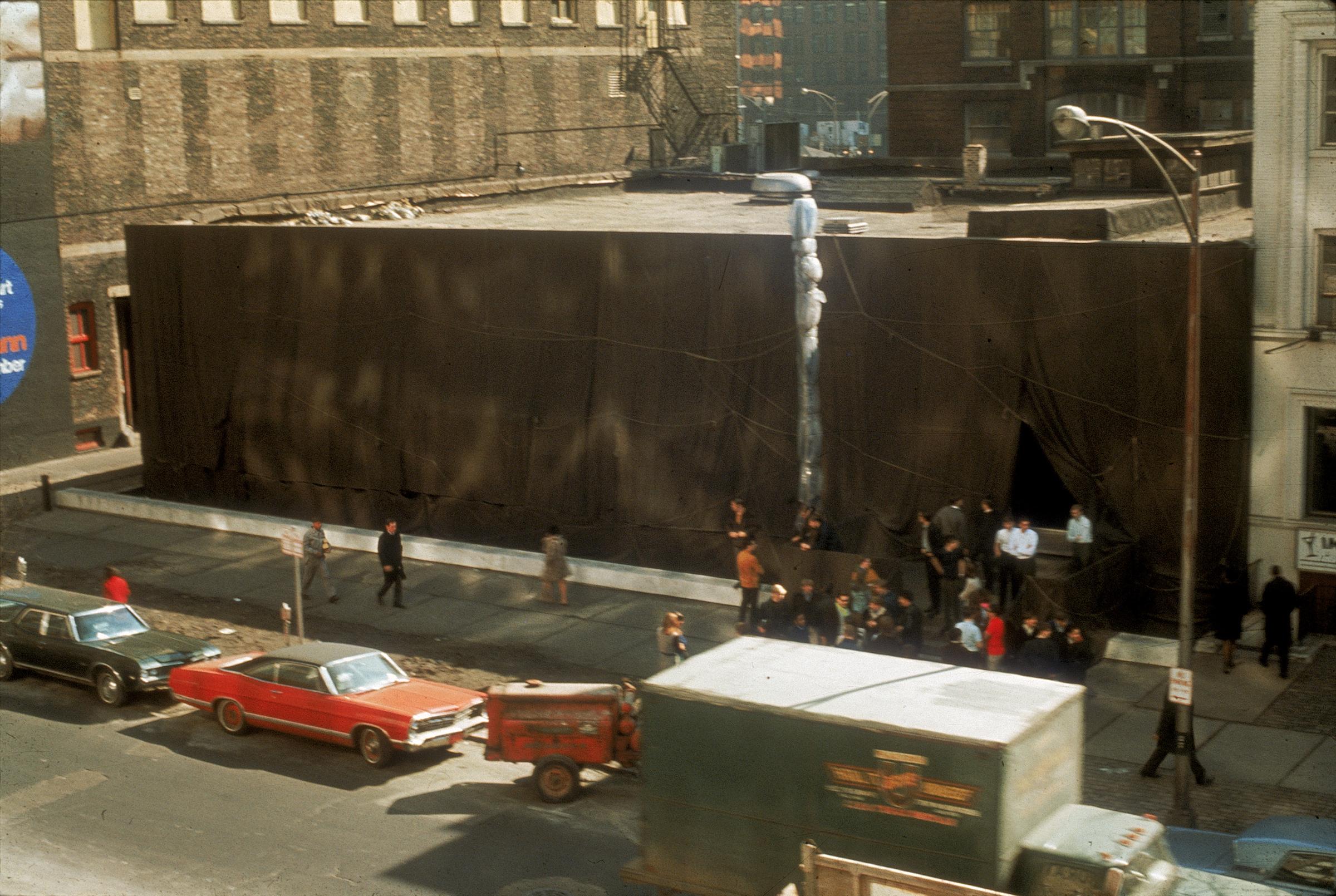 A street view photograph with people on the sidewalk looking at a building wrapped in fabric; cars are parked along the street in the foreground