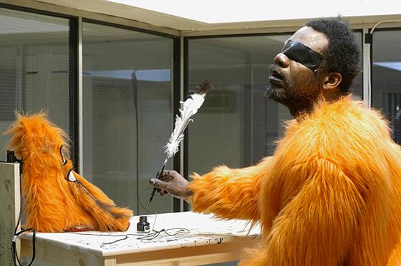 A man wearing a one-piece garment of bright orange fur sits at a desk in a glass room. He dips a quill into an inkwell. His eyes are taped shut with black duct tape; the headpiece for his orange suit is beside him on the desk.